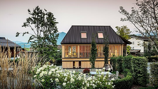Das „Badehaus“ des Hotels Hirschen in Schwarzenberg ist von einer liebevoll gestalteten Gartenlandschaft umgeben und liegt frontal vor einem romantischen Abendrot. Im Vordergrund blühen weiße Hortensien, Ziergräser und Büsche, die den offenen Außenbereich mit Sitzgruppe und Feuerstelle umrahmen. Im Zentrum steht das moderne Gebäude mit dem auffälligen PREFA-Stehfalzdach in der Farbe P.10 Nussbraun. Dessen präzise gearbeitete Scharen verlaufen schräg und verleihen dem Dach eine dynamische Wirkung. Die Fassade aus vertikalen, geschosshohen Holzlamellen verbindet moderne Formensprache mit regionalem Baustil. Im Hintergrund zeichnen sich die sanften Hügel und Berge des Bregenzerwaldes ab – ein harmonisches Zusammenspiel von Natur, Architektur und Tradition.