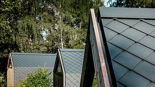 Plusieurs maisons minimalistes en forme de A du Secret Koi Garden s'harmonisent en bordure de forêt, près du lac Natterer. Leurs toits sont recouverts de petits panneaux d'aluminium PREFA en forme de losange, couleur P.10 anthracite, créant une texture saisissante évoquant des écailles de poisson. La surface mate dégage une impression de calme et d'élégance, tandis que l'aluminium PREFA, résistant aux intempéries et durable, offre une protection optimale en milieu alpin et marie à la perfection design et nature.