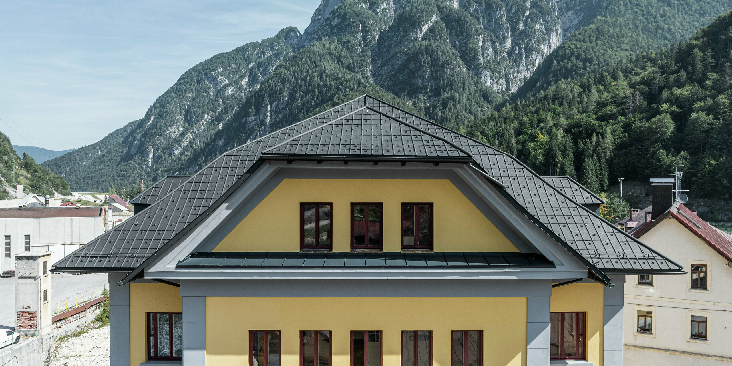 Nahaufnahme der Dacheindeckung des Museo Cave del Predil in Italien. Das gelbe Museumsgebäude wurde mit PREFA Dachplatten in P.10 Anthrazit eingedeckt. Die robuste Dachlösung aus Aluminium präsentiert sich harmonisch auf dem historischen Gebäude mit gelber Fassade. Im Hintergrund die beeindruckende Alpenlandschaft mit bewaldeten Berghängen, die eine extreme Witterungsbeständigkeit der PREFA Produkte erfordert.