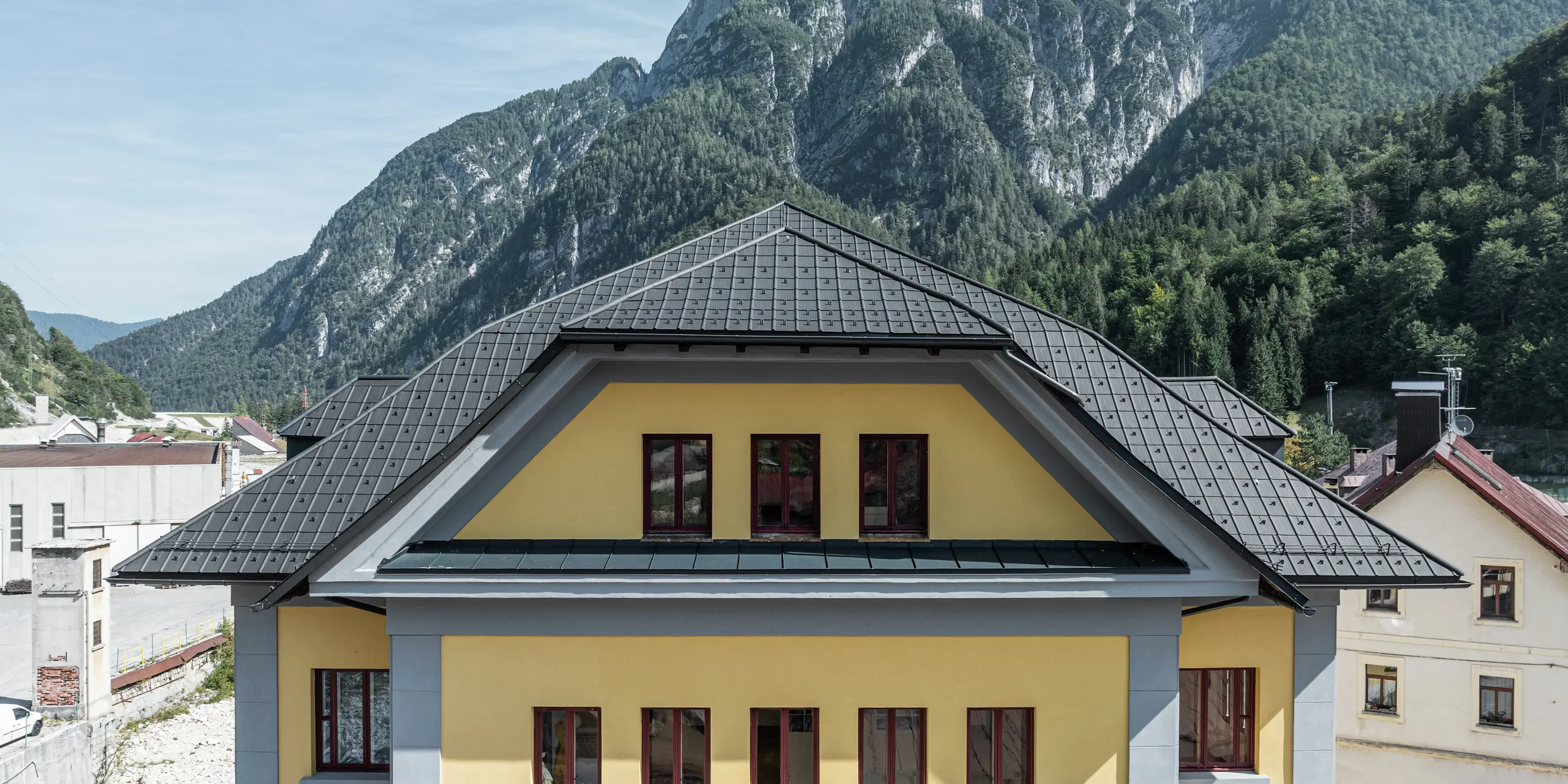 Gros plan de la couverture du Museo Cave del Predil en Italie. Le bâtiment jaune du musée a été recouvert de tuiles PREFA en P.10 anthracite. La solution de toiture robuste en aluminium s'intègre harmonieusement au bâtiment historique avec sa façade jaune. À l'arrière-plan se trouve l'impressionnant paysage alpin avec des pentes boisées, ce qui exige que les produits PREFA soient extrêmement résistants aux intempéries.
