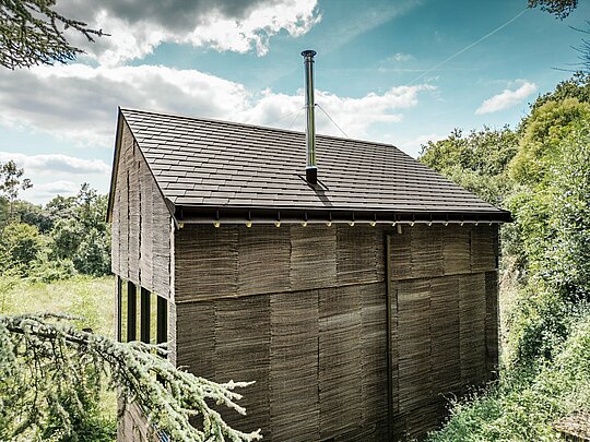 Vue arrière d'une maison avec des bardeaux de toiture PREFA en P.10 brun noisette et un système d'évacuation des eaux de pluie PREFA, entourée d'un terrain naturel dans l'ouest de la France.