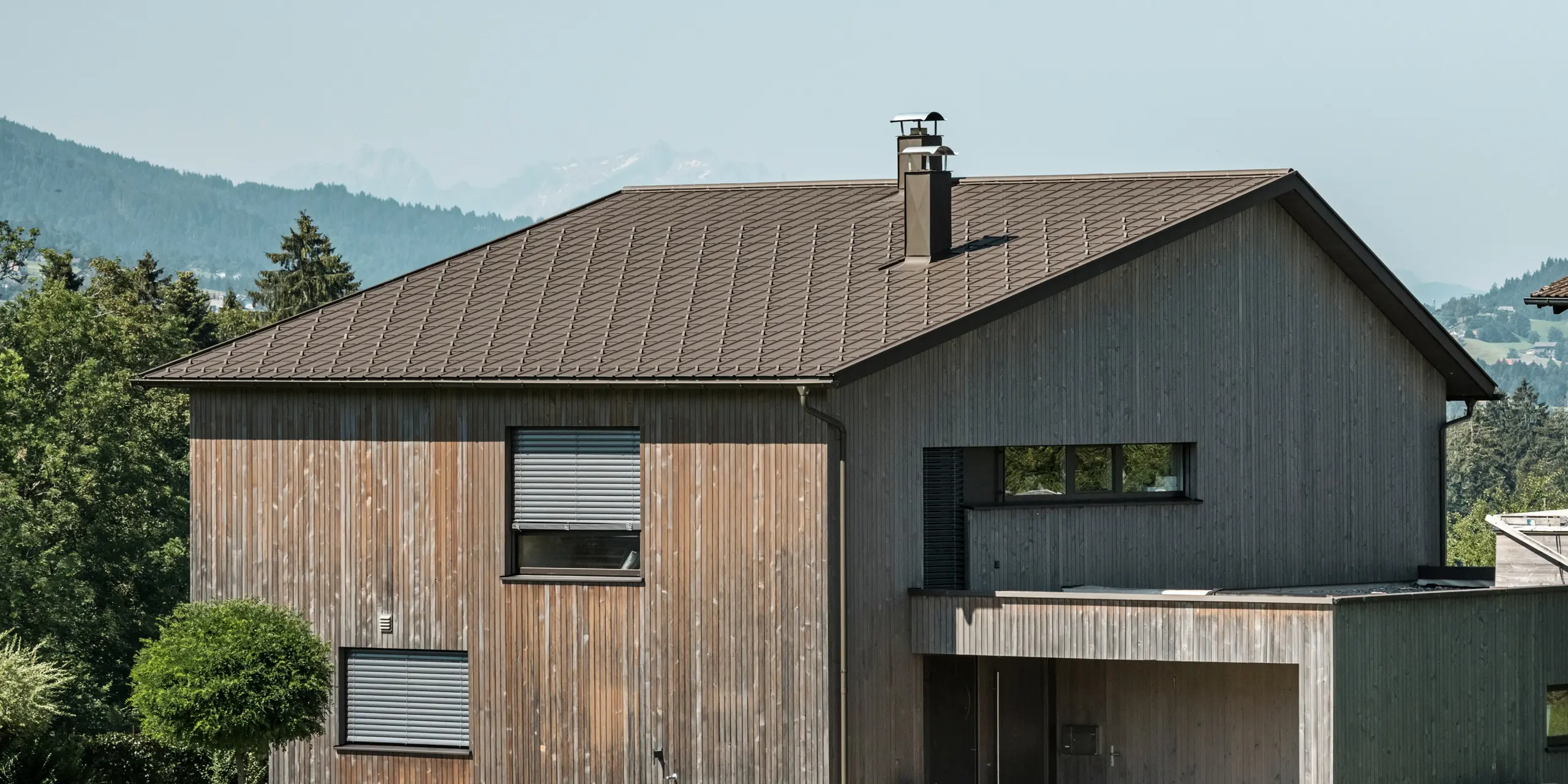 Vue latérale d'une maison individuelle contemporaine à Krumbach, avec une façade en bois naturel et une toiture en aluminium PREFA durable de couleur P.10 brun. L'harmonie des couleurs montre l'intégration élégante d'un design moderne et de matériaux traditionnels, tandis que les tuiles PREFA assurent une conception esthétique et fonctionnelle de la toiture.