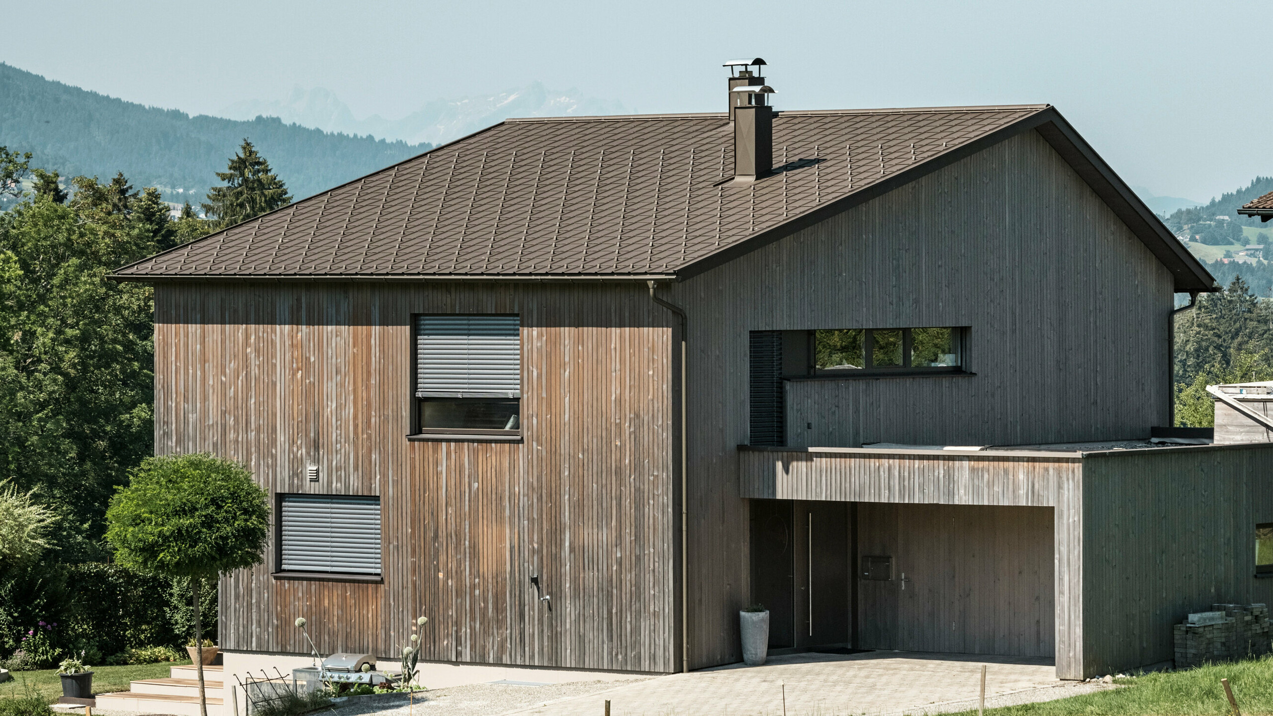 Seitenansicht eines zeitgenössischen Einfamilienhauses in Krumbach mit einer Kombination aus einer natürlichen Holzfassade und einem langlebigen PREFA Aluminiumdach in P.10 Braun. Die harmonische Farbgebung zeigt die elegante Integration von modernem Design und traditionellen Materialien, wobei die PREFA Dachplatten für eine ästhetische und funktionale Dachgestaltung sorgen.