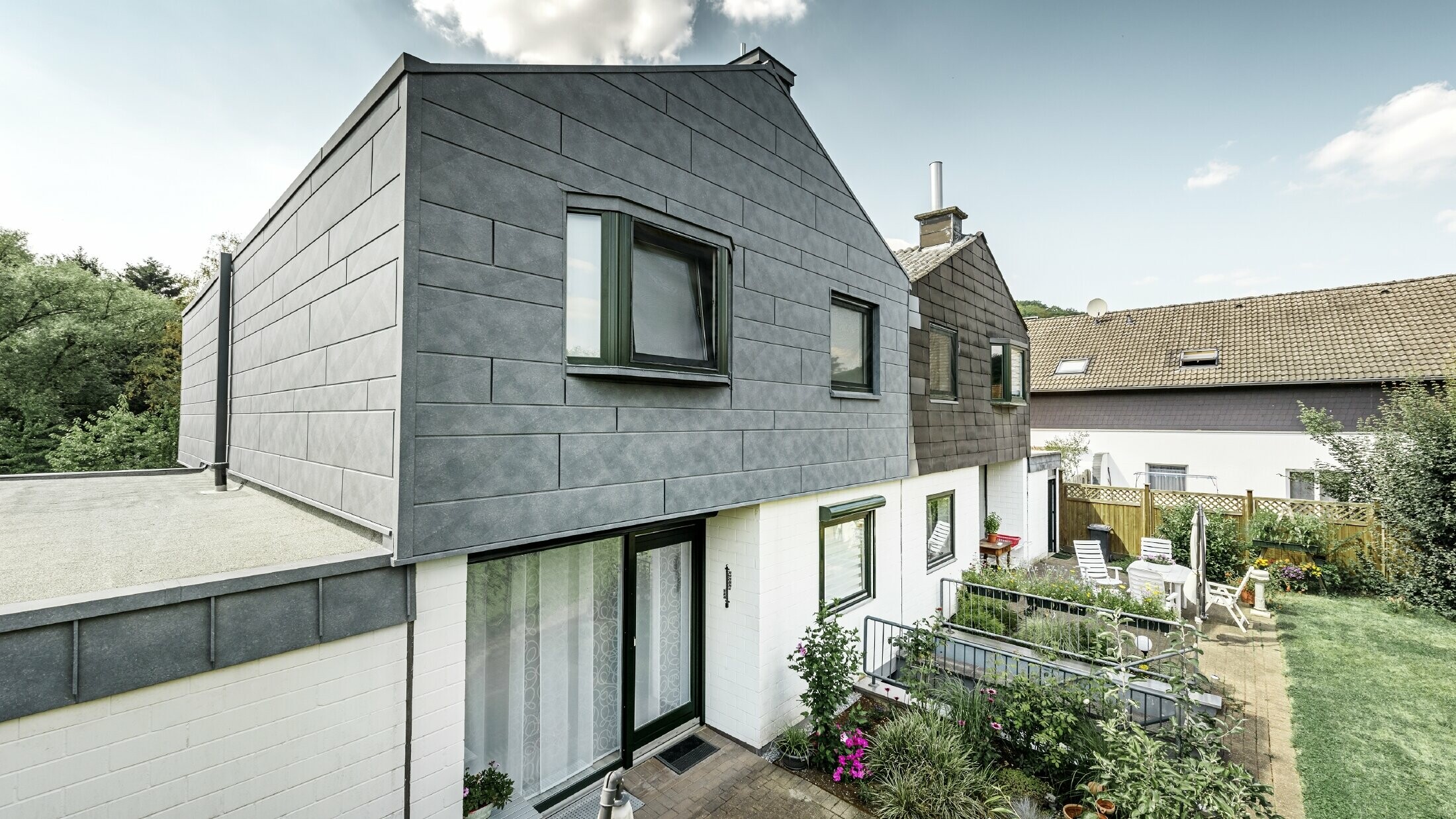 La partie haute du bâtiment de la moitié de maison jumelée a été rénovée avec l’élément de façade Siding.X PREFA en P.10 gris pierre. Juste à côté pour la comparaison, la moitié de maison avant la rénovation.