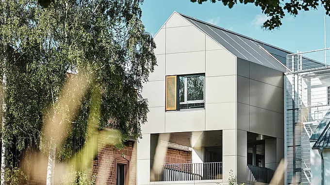 La photo montre la mairie moderne d'Alzenau, dont la façade est composée de panneaux composites en aluminium PREFA dans un élégant ton bronze. Le toit pointu et les grandes fenêtres soulignent le caractère contemporain du bâtiment. Au premier plan, des arbres et des espaces verts encadrent le bâtiment, tandis que l'éclairage doux et le ciel clair soulignent l'espace urbain moderne.