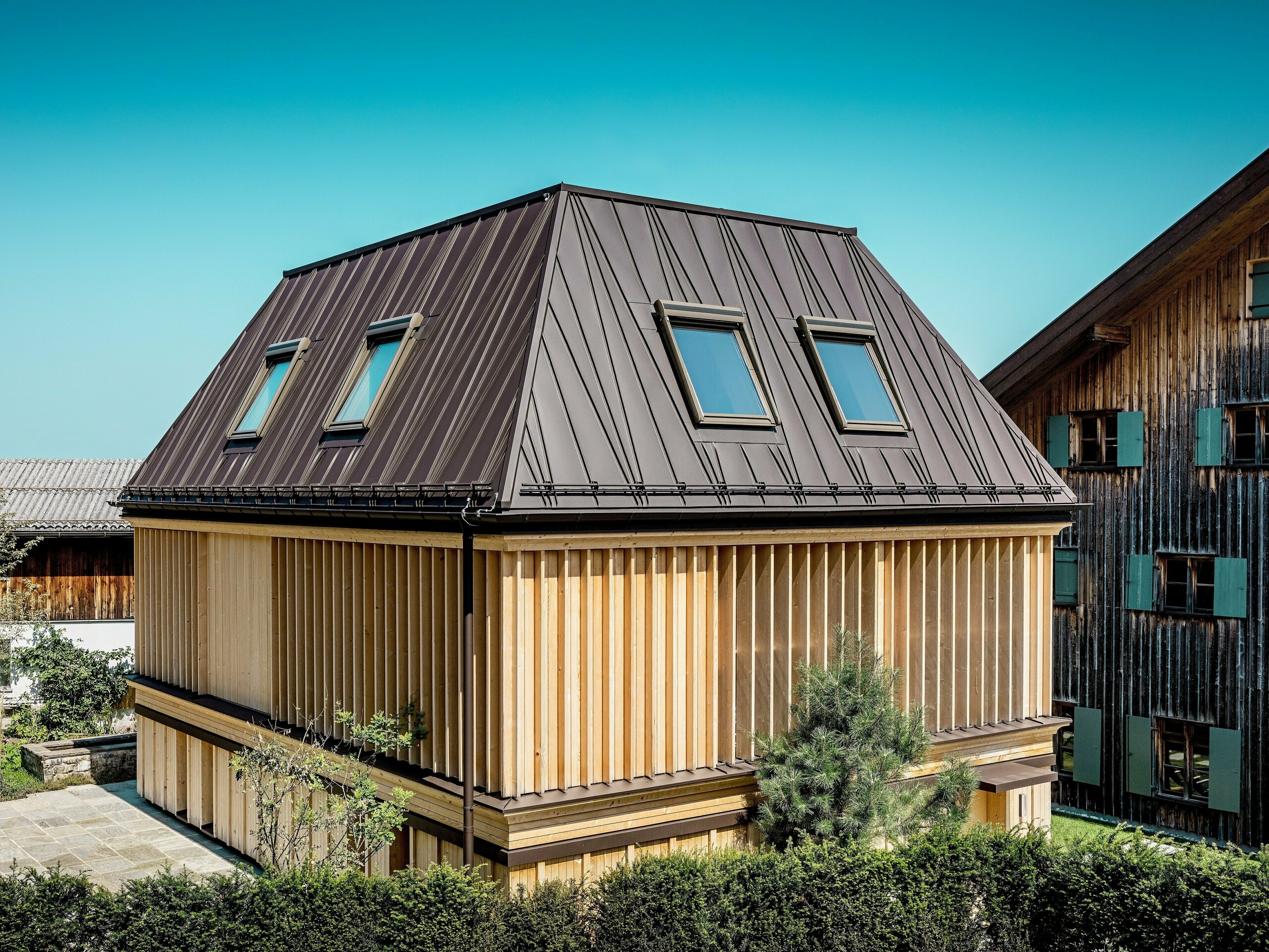 Das neu errichtete „Badehaus“ des Hotel Hirschen in Schwarzenberg bei Tageslicht. Im Vordergrund fällt das markante PREFA Stehfalzdach in P.10 Nussbraun auf, dessen atypisch gesetzte Falze dem Walmdach eine dynamische, moderne Anmutung verleihen. Die Dachneigung von 60 Grad unterstreicht die prägnante Architektur. Die Fassade besteht aus vertikal angeordneten, geschoßhohen Holzlamellen, die Licht und Schatten effektvoll inszenieren und zugleich einen funktionalen Sonnenschutz bieten. Das Zusammenspiel von traditioneller Bauform und innovativer Materialwahl schafft einen spannenden Kontrast zum denkmalgeschützten Bestandsgebäude im Hintergrund, das mit seiner verwitterten Holzfassade typisch für die Region Bregenzerwald ist.