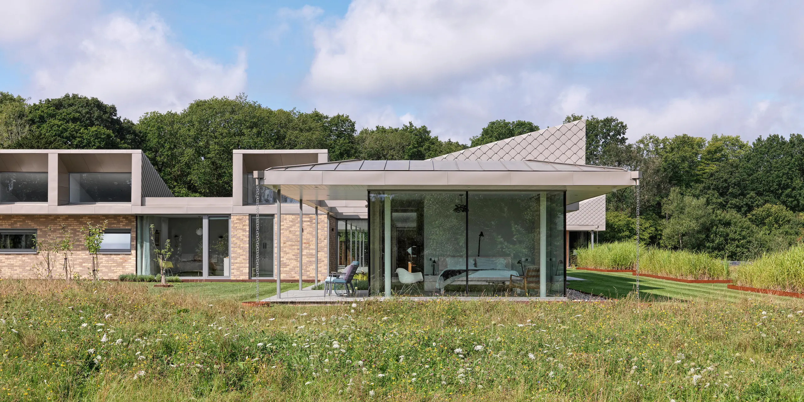 Vue latérale d'un bungalow moderne dans le West Sussex, doté d'une toiture en aluminium PREFALZ et de façades accentuées en losanges muraux PREFA 20 × 20 en P.10 bronze. Les baies vitrées des pièces à vivre s'ouvrent sur la prairie fleurie au premier plan, créant une connexion harmonieuse entre l'intérieur et la nature. Le système de toiture et de façade PREFA, résistant, assure non seulement la protection du bâtiment, mais lui confère également une élégance architecturale.