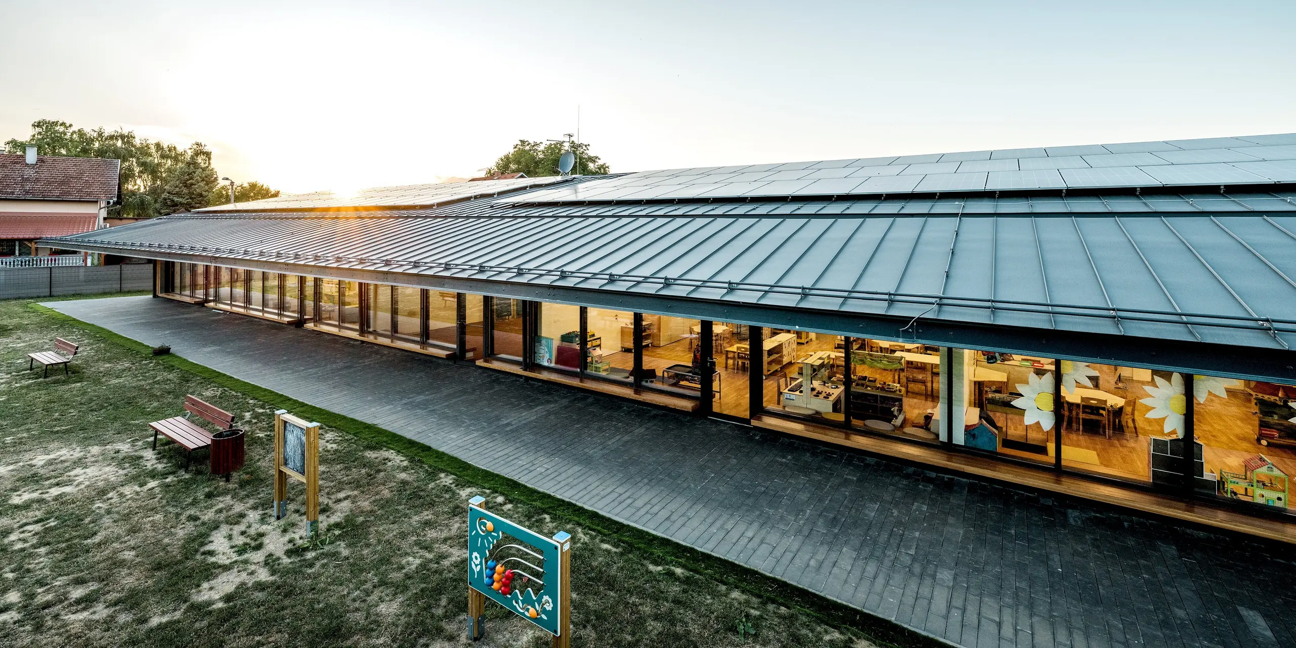 Moderner Kindergarten Svarozic mit weit auskragendem PREFALZ Aluminiumdach in P.10 Anthrazit, großflächigen Glasfronten und warmer Holzfassade. Die klare Dachstruktur prägt die Architektur und zeigt die langlebige, witterungsbeständige Qualität von PREFA Aluminium. Ein harmonisches Zusammenspiel aus Licht, Naturmaterialien und robustem Dachschutz schafft eine einladende Szene.