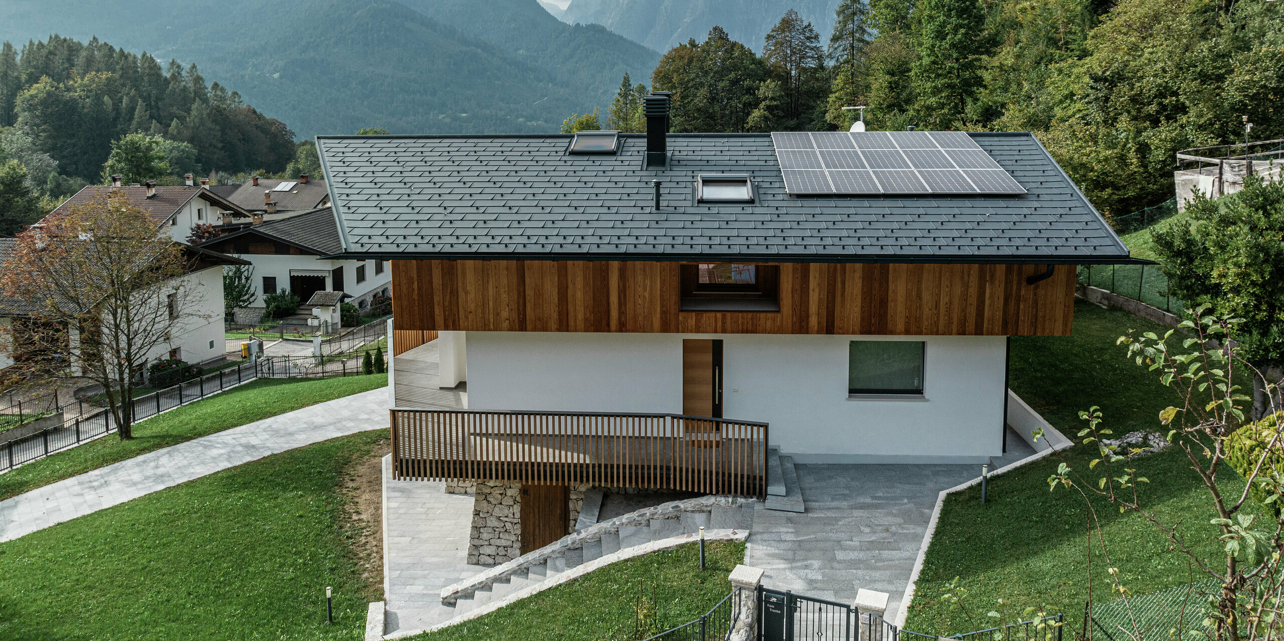 Seitenansicht eines modernen Einfamilienhauses in Agordo, Italien, mit PREFA Dachplatten R.16 in der Farbe P.10 Anthrazit. Das langlebige Aluminiumdach bietet optimalen Witterungsschutz und ist für die alpine Lage bestens geeignet. Eine PREFA Dachentwässerung mit Kastenrinne und Ablaufrohr sorgt für eine effiziente Wasserableitung. Die harmonische Kombination aus Holzfassade, weißer Putzfassade und nachhaltiger Photovoltaikanlage auf dem Dach unterstreicht das zeitlose Design des Gebäudes. Im Vordergrund ist der Eingangsbereich des Hauses zu sehen. Im Hintergrund sind ein bewölkter Himmel und viele Bäume zu erkennen. 