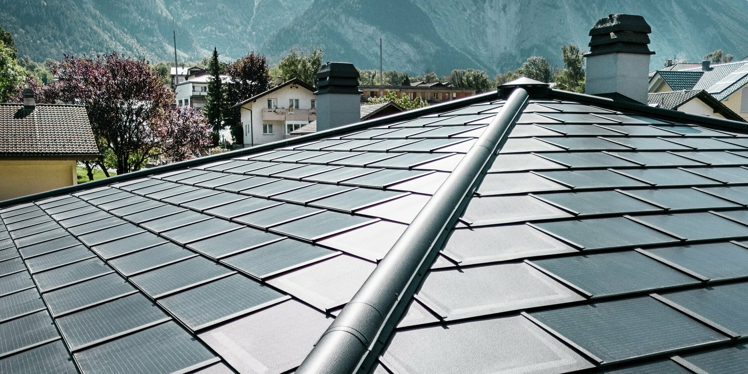 Die innovativen PREFA Solardachplatten in der Farbe P.10 Schwarz fügen sich perfekt in das Dachdesign der Weinkellerei Leukersonne in Susten ein. Das innovative Aluminiumdach mit integrierten Photovoltaikmodulen erzeugt umweltfreundlichen Solarstrom ohne optische Kompromisse. An den Rändern wurden die geradlinigen PREFA Dachplatten R.16 verlegt. Im Vordergrund des Bildes ist der präzise verarbeitete First des Zeltdaches zu sehen. Im Hintergrund sind weitere Dächer und der strahlend blaue Himmel zu erkennen. 