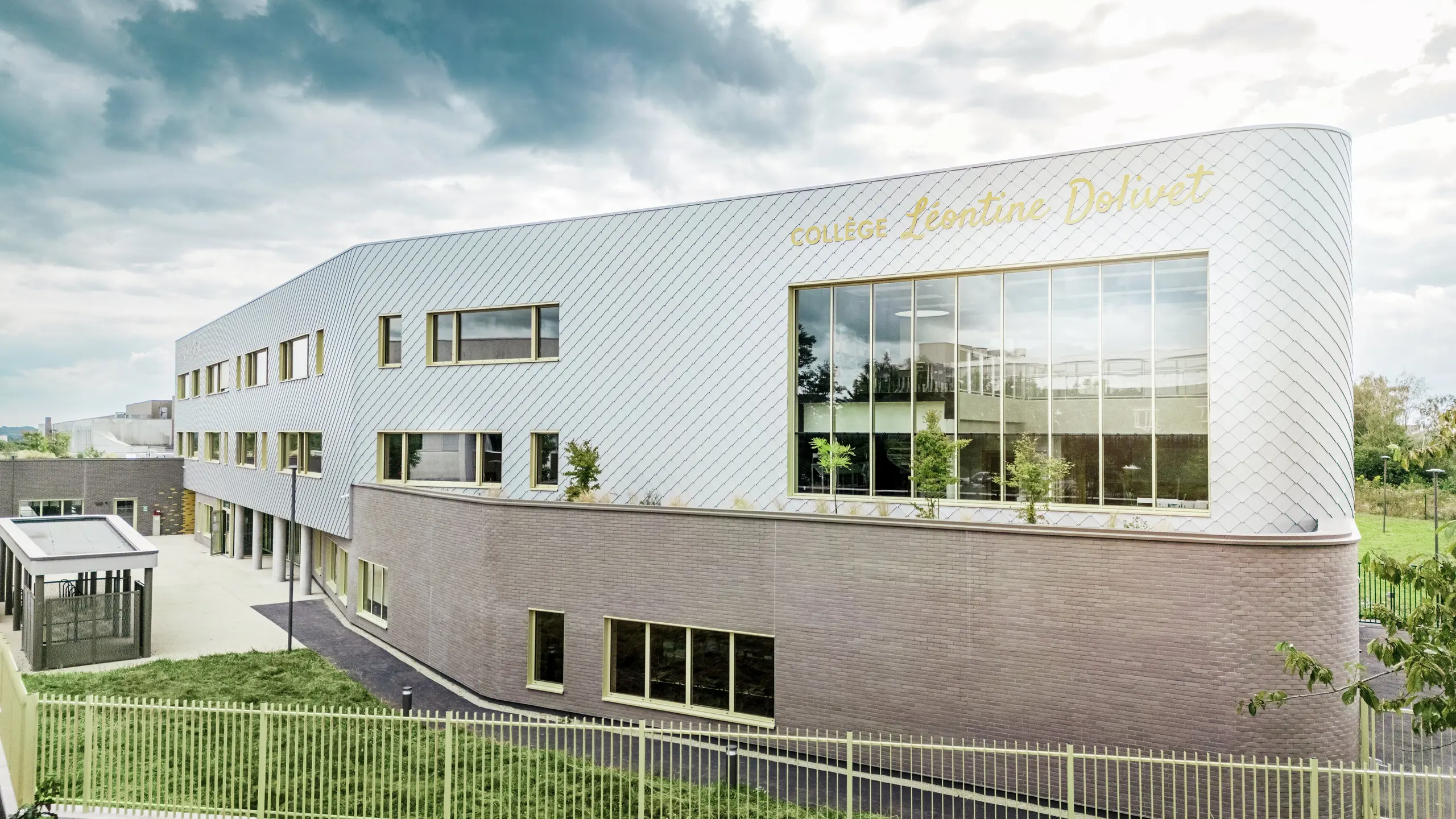 Vue latérale du Collège Léontine Dolivet à Cesson-Sévigné, dont la façade moderne en aluminium est composée de losanges PREFA argent métallisé. La structure diagonale crée un effet dynamique, tandis que les larges surfaces vitrées et les volets jaunes apportent des touches de couleur saisissantes. Au premier plan, des espaces verts et une clôture encadrent le bâtiment. Solution de façade durable, PREFA allie élégance, résistance aux intempéries et durabilité : une véritable prouesse architecturale aux lignes épurées et au métal scintillant.