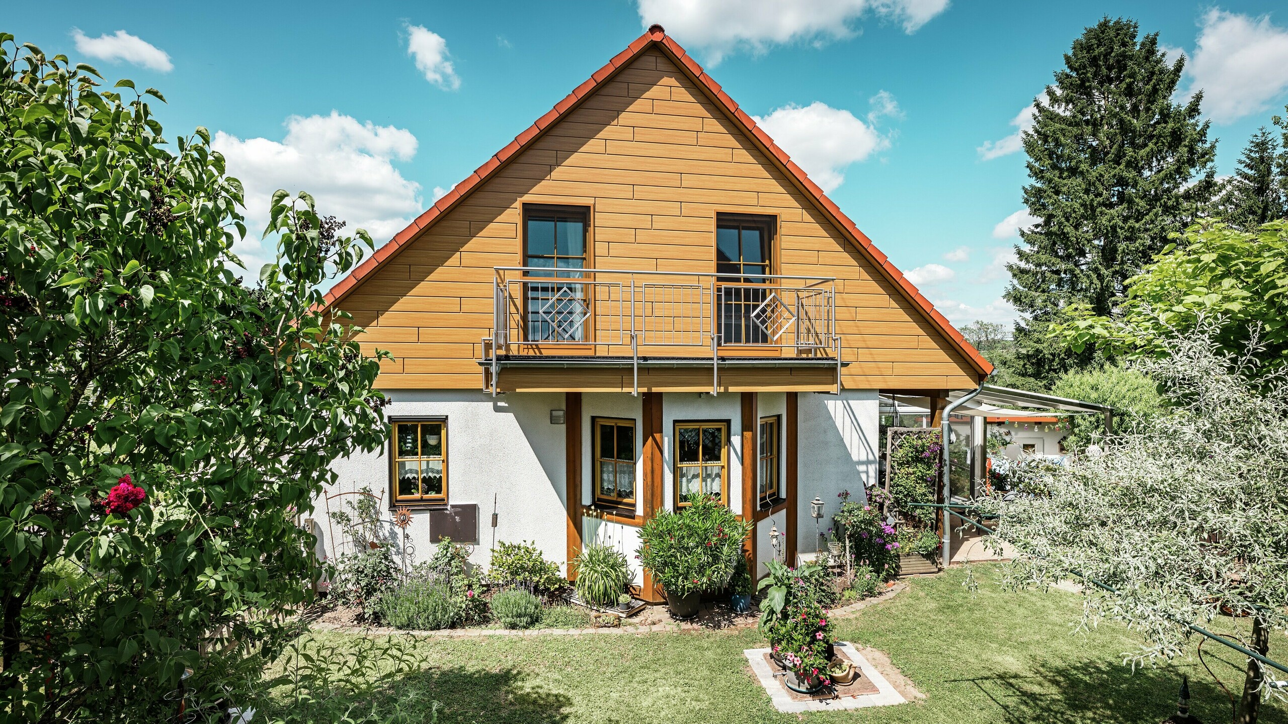 Sur cette image, le pignon rénové d'une maison individuelle à Blieskastel resplendit d'une chaleureuse teinte chêne naturel grâce au Siding PREFA. La façade en aluminium de haute qualité, à l'aspect bois réaliste, crée un contraste saisissant et s'harmonise parfaitement avec le toit de tuiles rouges et le jardin luxuriant. Entourée de fleurs, d'arbustes et de verdure, la maison paraît accueillante et impeccablement entretenue. La surface PREFA allie un aspect naturel à des avantages fonctionnels tels qu'une grande résistance aux intempéries, une stabilité optimale et une durabilité quasi sans entretien – un atout esthétique indéniable et une valeur ajoutée durable.