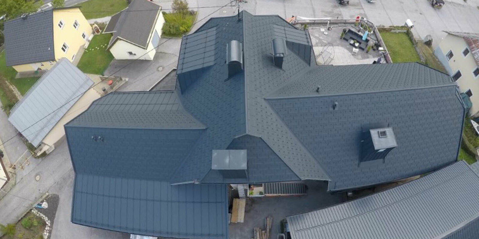 Rénovation d’une grande surface de toiture présentant de nombreux détails : noues, pignons et cheminées. Le toit est recouvert de tuiles R.16 en aluminium de PREFA, couleur anthracite.