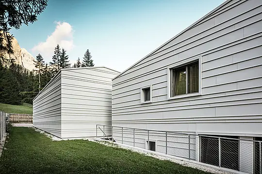 La face envers de la caserne « Tempesti » à Corvara offre une perspective saisissante : des arbres, un ciel bleu éclatant et des montagnes mises en valeur par le soleil. La façade claire en aluminium, avec sa structure nette, montre le raffinement technique de PREFALZ dans la couleur P.10 blanc Prefa. Les lames horizontales en aluminium reflètent la lumière, offrent une protection optimale contre les intempéries et s'intègrent harmonieusement dans l'environnement alpin. Un exemple parfait d'architecture militaire moderne et durable !