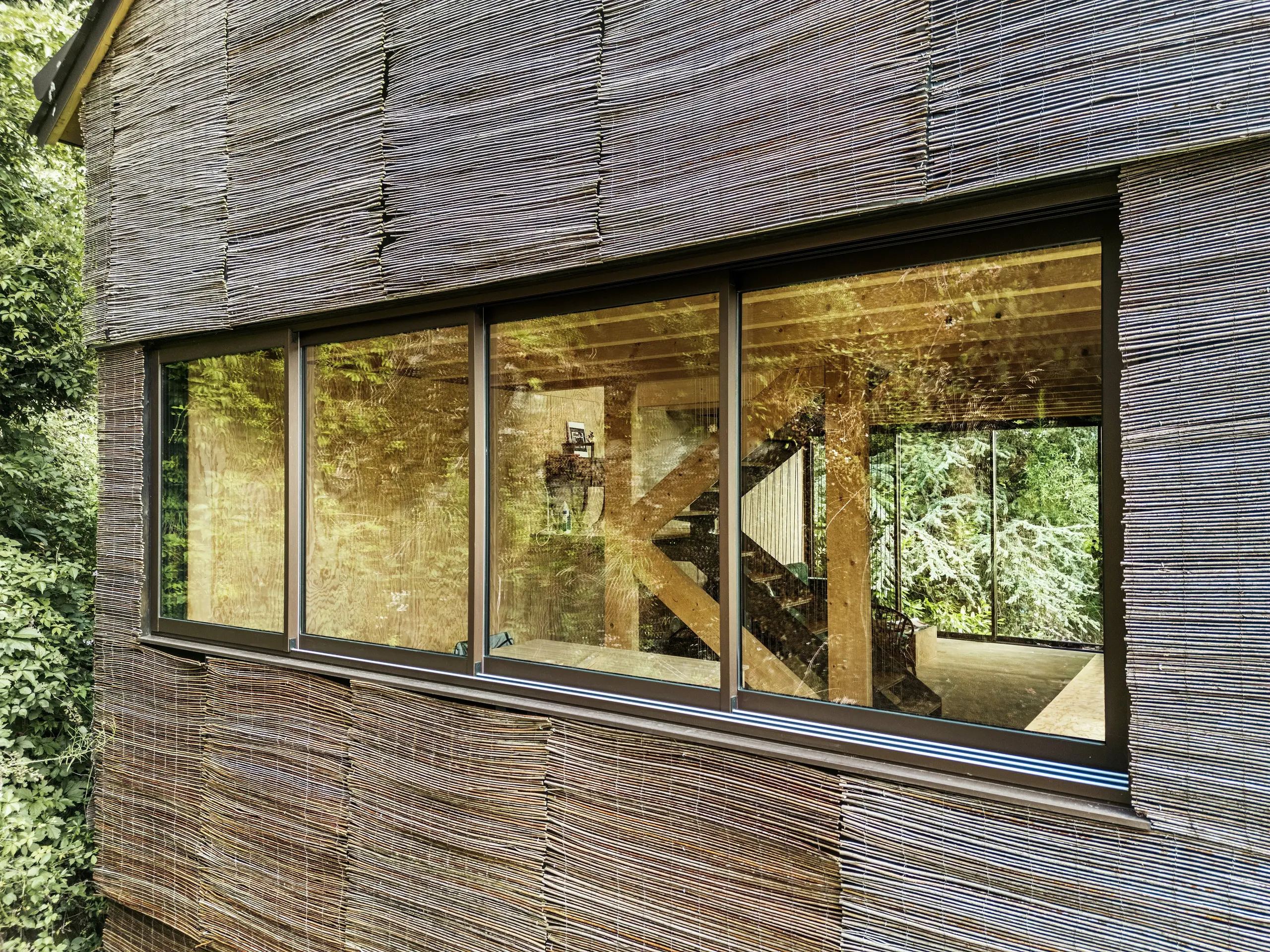 Détail d'une fenêtre avec un cadre en aluminium moderne et une façade en bois de saule sur une maison en France – conception durable avec des bardeaux de toiture PREFA et l'environnement naturel.