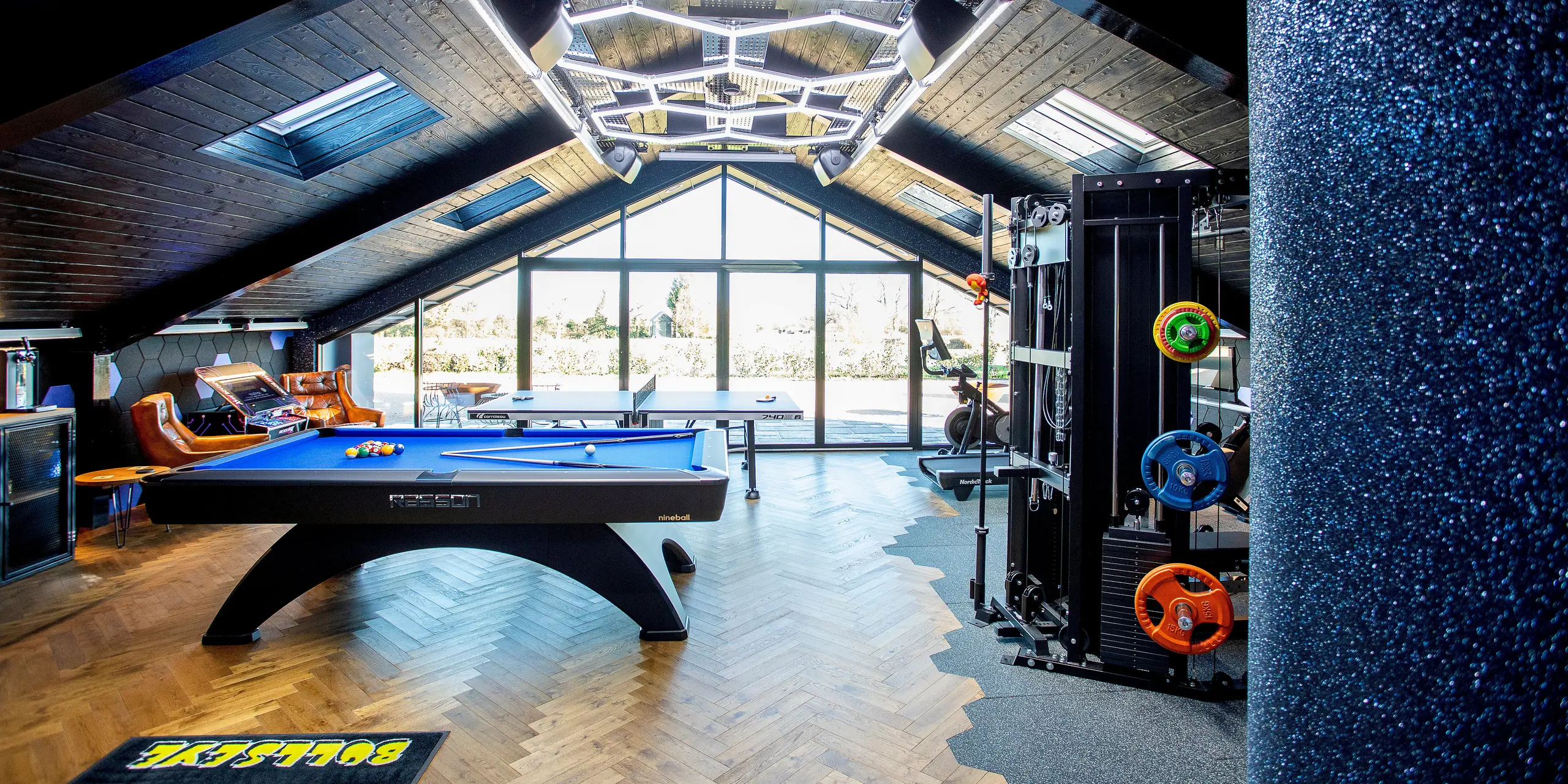 Vue intérieure d'un bâtiment de loisirs moderne, doté d'une large façade vitrée et d'une toiture à l'architecture remarquable. Cet espace polyvalent comprend un billard, des appareils de fitness, une table de ping-pong et une salle de jeux. Des matériaux de qualité, un éclairage dynamique et un parquet créent une ambiance chaleureuse et stimulante.