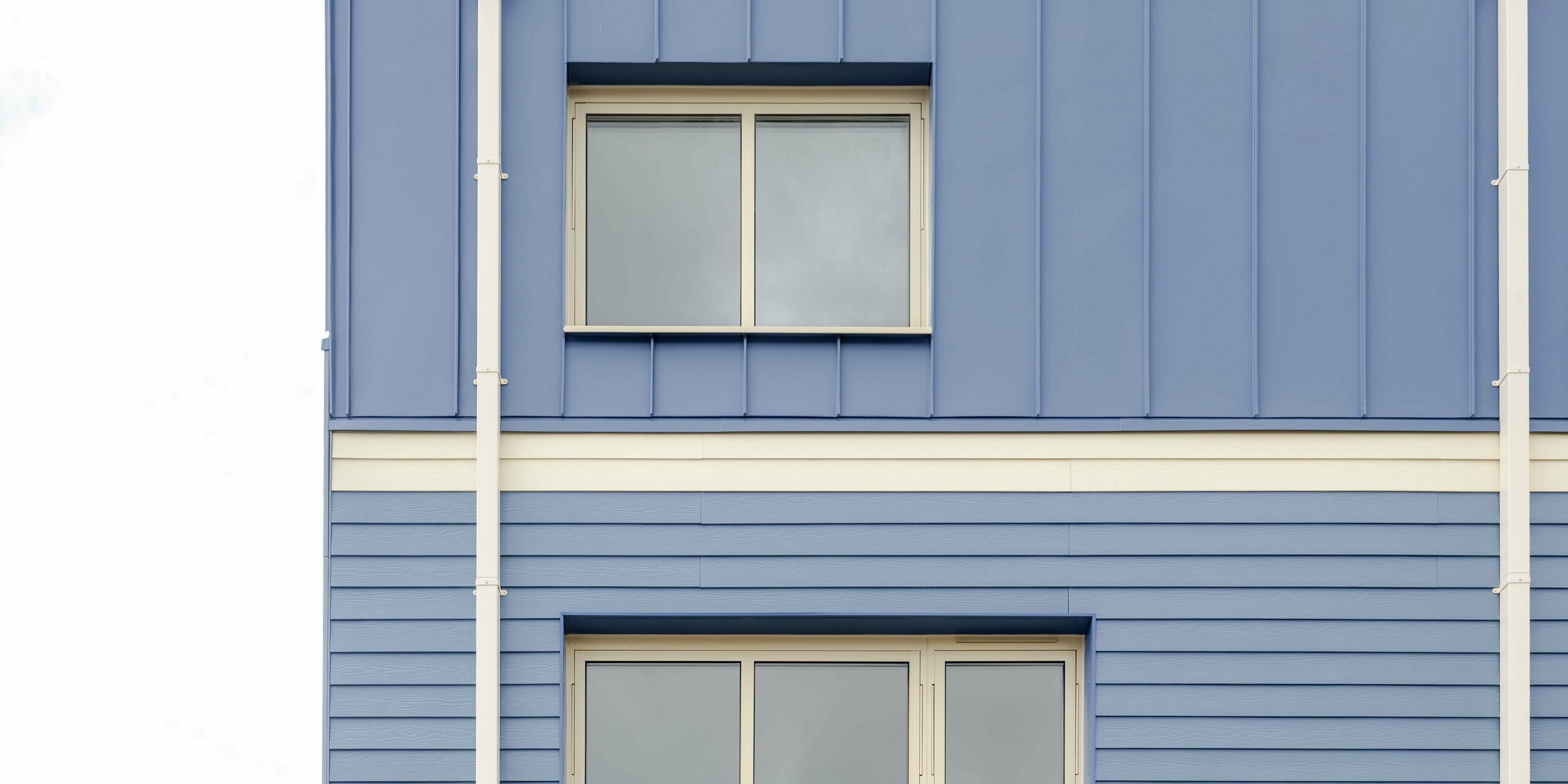 Die Aufnahme zeigt die Rückseite eines Goldsmith‑Mews‑Reihenhauses mit der markanten PREFA FALZONAL Verkleidung in Taubenblau. Die klar strukturierten, vertikalen Aluminiumfalze im oberen Bereich treffen auf die horizontalen Paneele des Mittelgeschosses und erzeugen ein stark gegliedertes Fassadenbild. Die hellen unteren Bereiche mit großen Terrassentüren öffnen die Architektur zum Garten. Das hochwertige PREFA Aluminium bietet hohe Farb- und Formstabilität, dauerhaften Witterungsschutz und ermöglicht eine moderne, präzise gestaltete Gebäudehülle mit klarer Wiedererkennung.