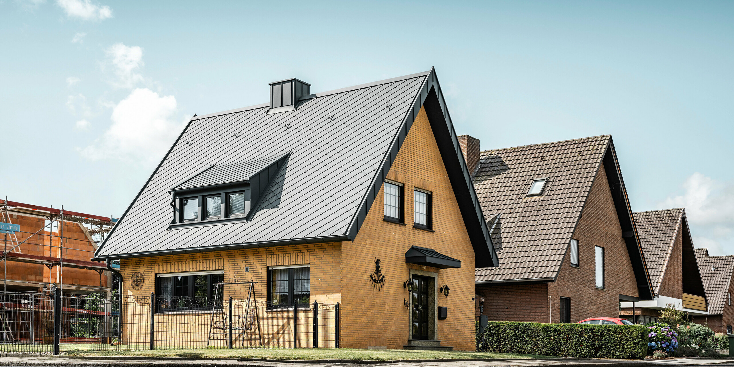 Ein traditionelles gelbes Backsteinhaus in Tönisvorst mit einem spitzen Giebeldach, bedeckt mit anthrazitfarbenen PREFA Dachrauten 29×29. Die Schleppgaube und der Schornstein, auch in Anthrazit gehalten, fügen sich in das Dach ein. Analog dazu runden die Regenrinne und das Ablaufrohr das Erscheinungsbild ab. Das Haus präsentiert sich in einer Wohnsiedlung, umgeben von ähnlichen Gebäuden, wobei sein Dach für einen modernen Akzent sorgt.