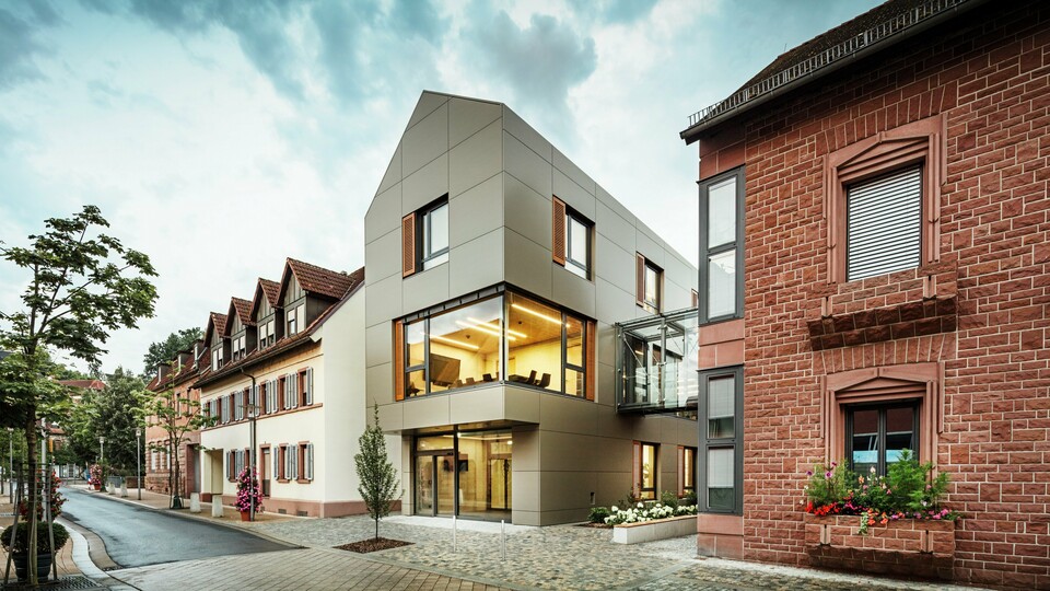 Das Bild zeigt das moderne Rathaus in Alzenau, das mit Aluminium-Verbundplatten von PREFA in einem warmen Bronze-Ton verkleidet ist. Die Fassade wird durch große Fensterflächen unterbrochen, die einen Blick auf den Innenraum gewähren, der durch warmes Licht erleuchtet wird. Der gläserne Verbindungsgang im rechten Teil des Bildes verbindet das Gebäude mit einem benachbarten, traditionellen Gebäude aus Ziegelstein, was einen spannenden Kontrast zwischen moderner und historischer Architektur schafft. 