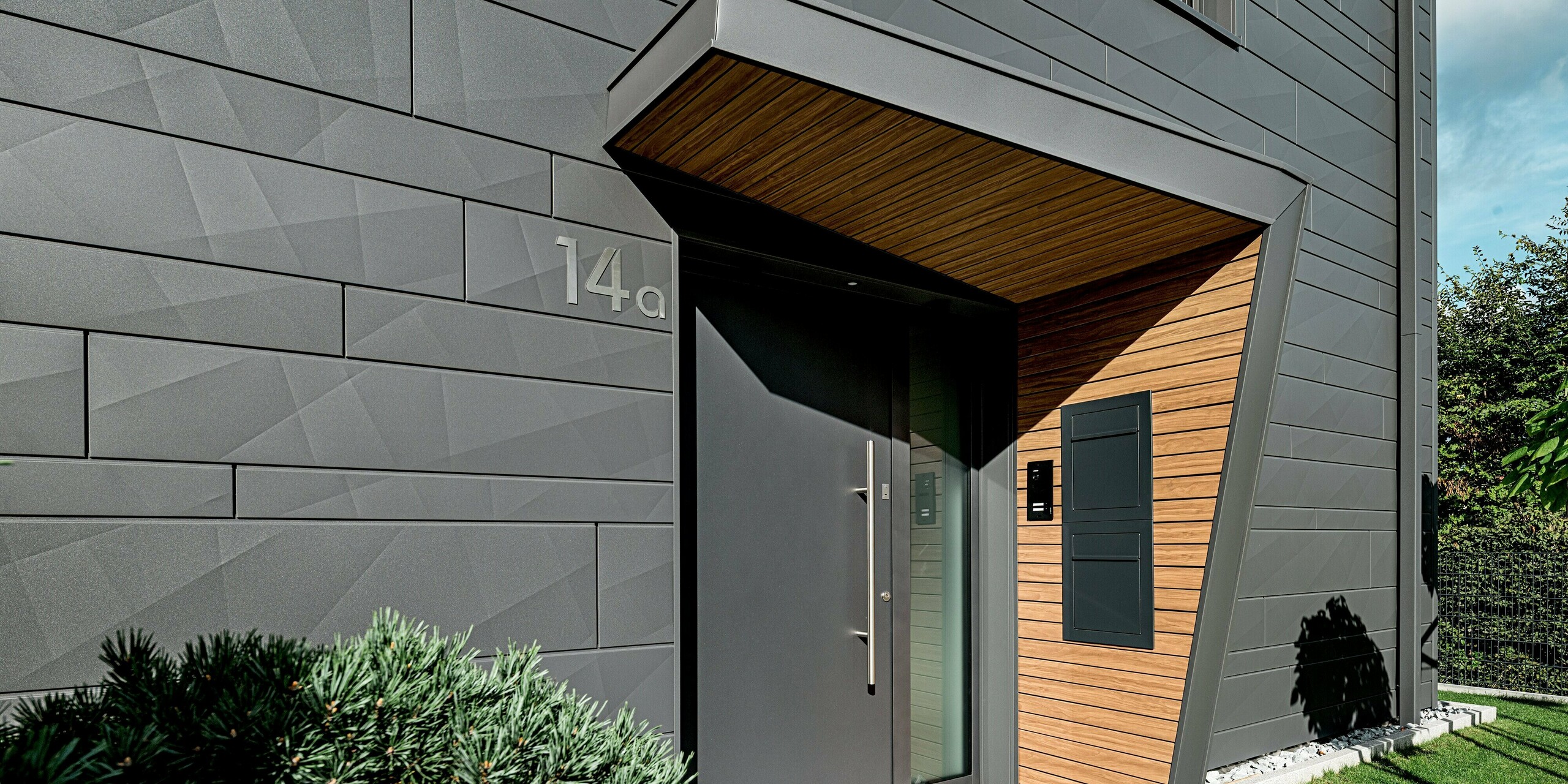Detailaufnahme des Eingangs eines modernen Einfamilienhauses in Heidenheim mit einer präzise verarbeiteten PREFA Siding.X Fassade in P.10 Dunkelgrau. Die markanten Kantungen und unterschiedlichen Paneelbreiten erzeugen ein dynamisches Lichtspiel. Der tief eingeschnittene, holzverkleidete Eingangsbereich bildet einen warmen Kontrast zur robusten Aluminiumoberfläche. Die klar gestaltete Haustüre, der strukturierte Schattenwurf und die umliegende Bepflanzung betonen die hochwertige, witterungsbeständige Bauweise und den modernen Charakter der PREFA Fassadenlösung.