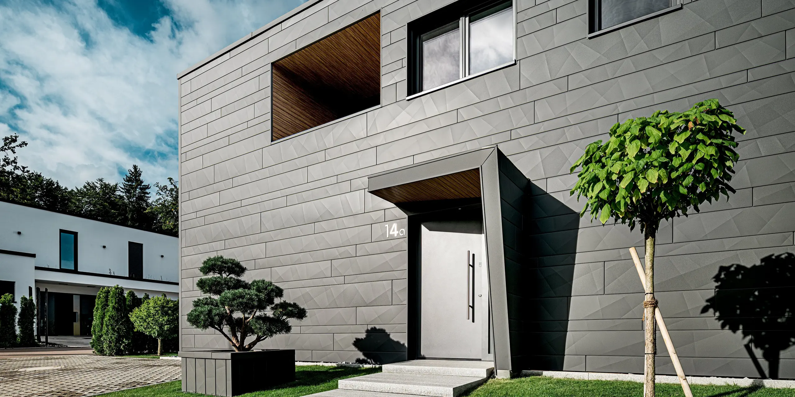 Cette maison individuelle moderne à Heidenheim présente une façade Siding.X PREFA remarquable, couleur P.10 gris sombre, dont la structure angulaire souligne la pureté du design. L'entrée, avec son renfoncement profond revêtu de bois, crée un point focal architectural captivant. Au premier plan, un topiaire taillé et un jeune arbre en forme de globe apportent des touches de dynamisme. La façade en aluminium durable et les tuyaux de descente carrés PREFA intégrés garantissent une protection durable, un style contemporain et une apparence générale de grande qualité.