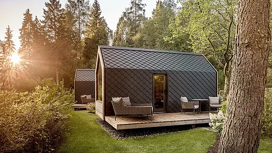 Nichée dans un écrin de verdure paisible au bord du lac Natterer, une minuscule maison du Secret Koi Garden est baignée par la douce lumière du soleil couchant. Son toit et sa façade sont revêtus de petits panneaux d'aluminium PREFA en forme de losange, couleur P.10 anthracite, dont la texture évoque la peau d'un poisson. Robuste et durable, la structure en aluminium offre une protection efficace contre les intempéries et souligne l'architecture épurée et intemporelle de ce glamping en pleine nature.