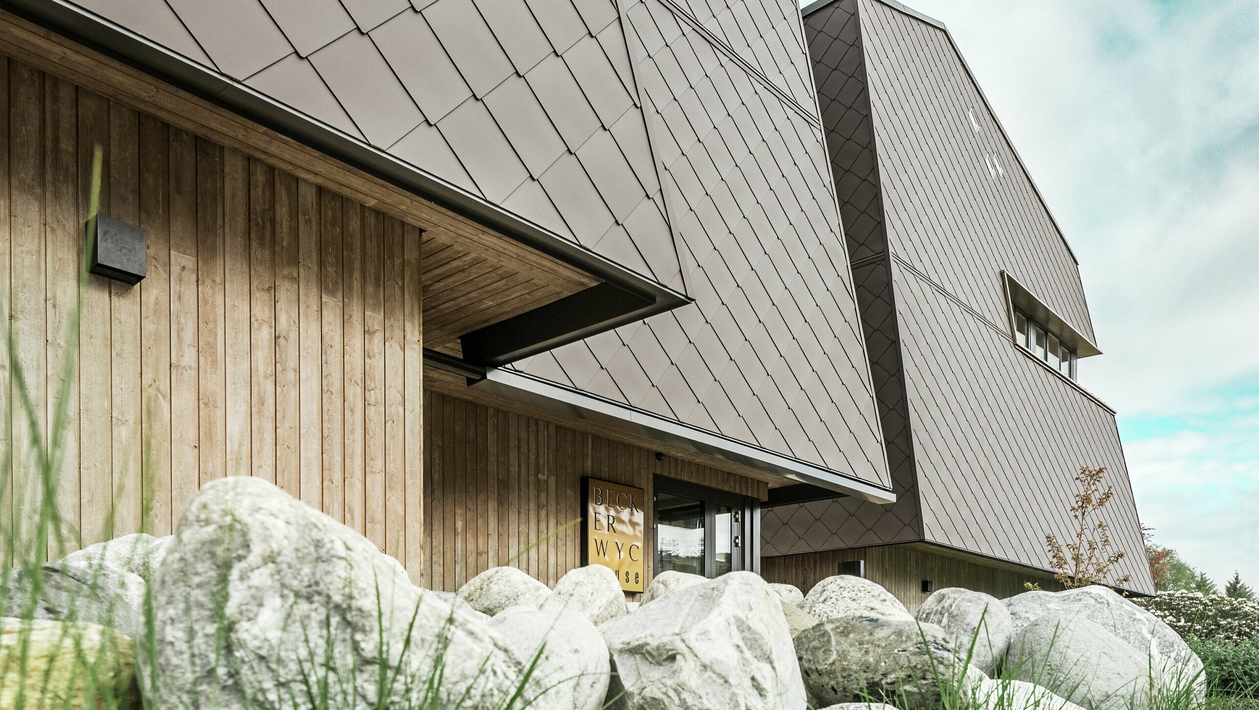 Vue latérale d'un hôtel, la façade du rez-de-chaussée est revêtue verticalement de bois plutôt sombre. Au-dessus se trouve un avant-toit, à partir duquel la façade est revêtue de plaques d'aluminium brun noisette.