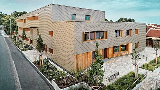 La lumière du soir baigne la crèche d'Alzenau d'une douce lueur, mettant en valeur sa façade de haute qualité, réalisée en losanges de façade 44x44 PREFA couleur bronze. L'alliance de la façade structurée en aluminium et des surfaces verticales en bois crée un jeu dynamique de matières, de couleurs et de textures. De larges fenêtres inondent les intérieurs de lumière, tandis que les espaces verts environnants et le parvis pavé invitent à la flânerie. Cette image illustre avec brio comment l'architecture contemporaine, la fonctionnalité et le développement durable s'intègrent harmonieusement aux produits PREFA, en plein cœur du tissu urbain d'Alzenau.