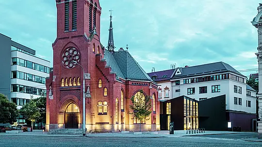 L'image illustre la fusion harmonieuse de l'architecture historique et moderne à Olomouc. L'imposante église rouge, avec sa flèche caractéristique et ses fenêtres gothiques, constitue le cœur historique de l'ensemble. Juste à côté s'élève le nouveau bâtiment moderne, dont la façade en panneaux composites en aluminium mat PREFABOND et en verre crée un contraste élégant avec l'architecture traditionnelle. Les larges façades vitrées offrent des aperçus sur le nouveau bâtiment, relié de manière innovante à l'église. Par sa forme précise et ses lignes épurées, la structure moderne contribue à l'atmosphère contemporaine sans compromettre le caractère historique de l'édifice. Cette symbiose réussie démontre comment des matériaux de haute qualité comme PREFABOND constituent le socle idéal pour la fusion de la tradition et de la modernité.