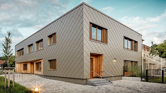 La crèche moderne d'Alzenau séduit par ses lignes épurées et sa façade en aluminium PREFA couleur bronze de haute qualité. Composée de losange de façade PREFA de 44 x 44 cm, cette façade confère au bâtiment une allure élégante et structurée, unifiant visuellement les espaces fonctionnels en un ensemble harmonieux. Le bardage en bois aux entrées offre un contraste chaleureux et naturel avec la robustesse de l'aluminium. L'alliance d'une construction solide, d'éléments en bois et d'une façade ventilée à double paroi illustre parfaitement les pratiques de construction durable. L'enveloppe du bâtiment PREFA, nécessitant peu d'entretien, est non seulement recyclable, mais aussi extrêmement résistante aux intempéries – un atout majeur pour les établissements scolaires d'avant-garde répondant à des exigences architecturales élevées.