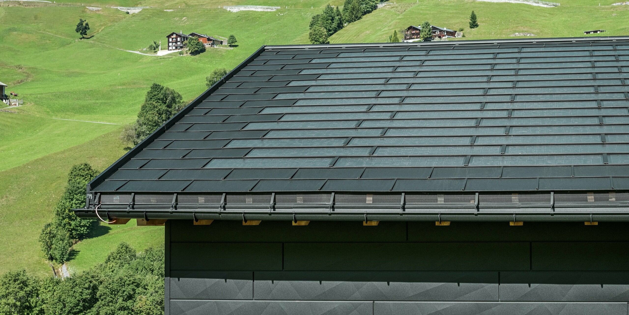Gros plan sur l'avant-toit d'une maison familiale moderne dans les environs alpins d'Ascharina, en Suisse. Le toit a été recouvert de la tuile solaire innovante PREFA et du R.16 traditionnel, tous deux en P.10 noir. La gouttière suspendue PREFA et le tuyau de descente PREFA assurent un drainage fiable du toit. Les panneaux de façade distinctifs PREFA Siding.X sont visibles au bas de l'image. Les lignes claires des produits en aluminium soulignent le design moderne au milieu d'un paysage de montagne pittoresque.