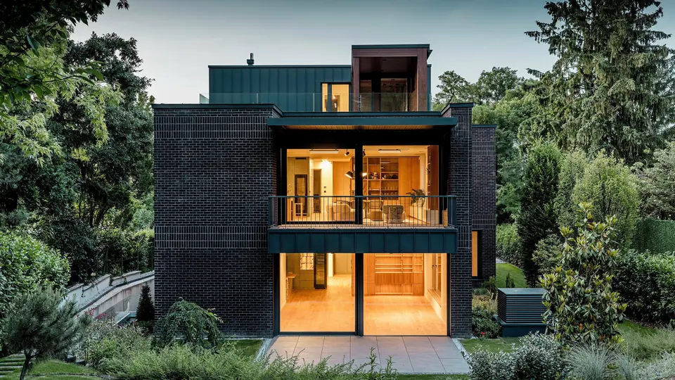 La photographie dévoile la façade arrière moderne de cette villa multigénérationnelle à Budapest, au crépuscule. De larges baies vitrées ouvrent sur des intérieurs baignés d'une lumière chaleureuse, tandis que l'alliance de la brique rouge foncé et de l'aluminium PREFALZ P.10 anthracite souligne la pureté des lignes architecturales. La précision de l'exécution des joints debout angulaires témoigne de la qualité de fabrication et de la durabilité des produits en aluminium PREFA. Entouré d'une végétation luxuriante, le bâtiment s'intègre harmonieusement au jardin et affirme un caractère élégant et contemporain.