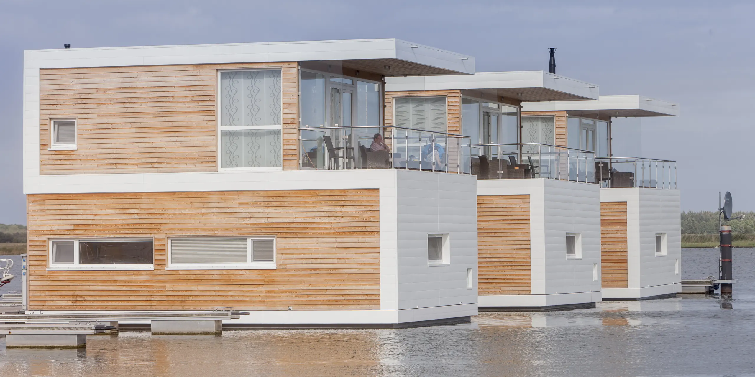 Vor dramatischer Wolkenkulisse präsentieren sich die schwimmenden Häuser in Stralsund als architektonischer Blickfang auf dem Wasser. Die Kombination aus warmen Holzfassaden, großzügigen Glasfronten und PREFA Sidings in P.10 Prefaweiß verleiht den Floating Homes eine moderne, maritime Ausstrahlung. Die langlebigen Aluminiumfassaden überzeugen durch Korrosionsbeständigkeit, Witterungsfestigkeit und eine dauerhaft hochwertige Optik im anspruchsvollen Hafenumfeld.
