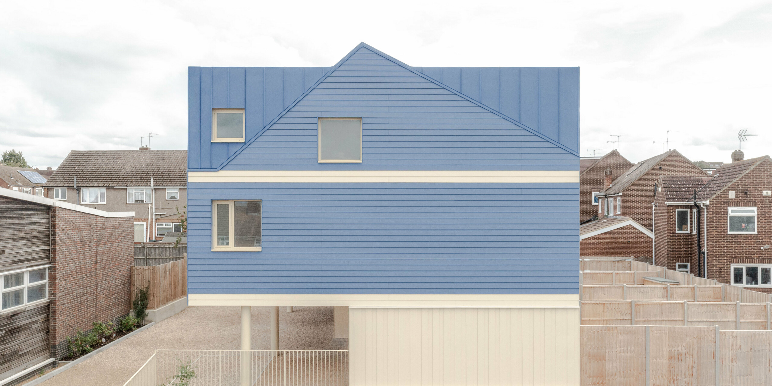 Das Bild zeigt die markante, taubenblaue FALZONAL Dach‑ und Fassadenverkleidung der Goldsmith Mews Reihenhausanlage, die sich klar von der umliegenden Bebauung abhebt. Die präzisen Falzstrukturen verleihen der Aluminiumoberfläche Tiefe und Rhythmus, während die hellen Erdgeschossbereiche einen starken, modernen Kontrast schaffen. Die aufgeständerte Bauweise wirkt leicht und offen. Das robuste, farbstabile PREFA Aluminium bietet langlebigen Witterungsschutz, hohe Formbeständigkeit und ermöglicht eine klare, mutige Architektur, die dem Gebäudeensemble einen unverwechselbaren Charakter verleiht.