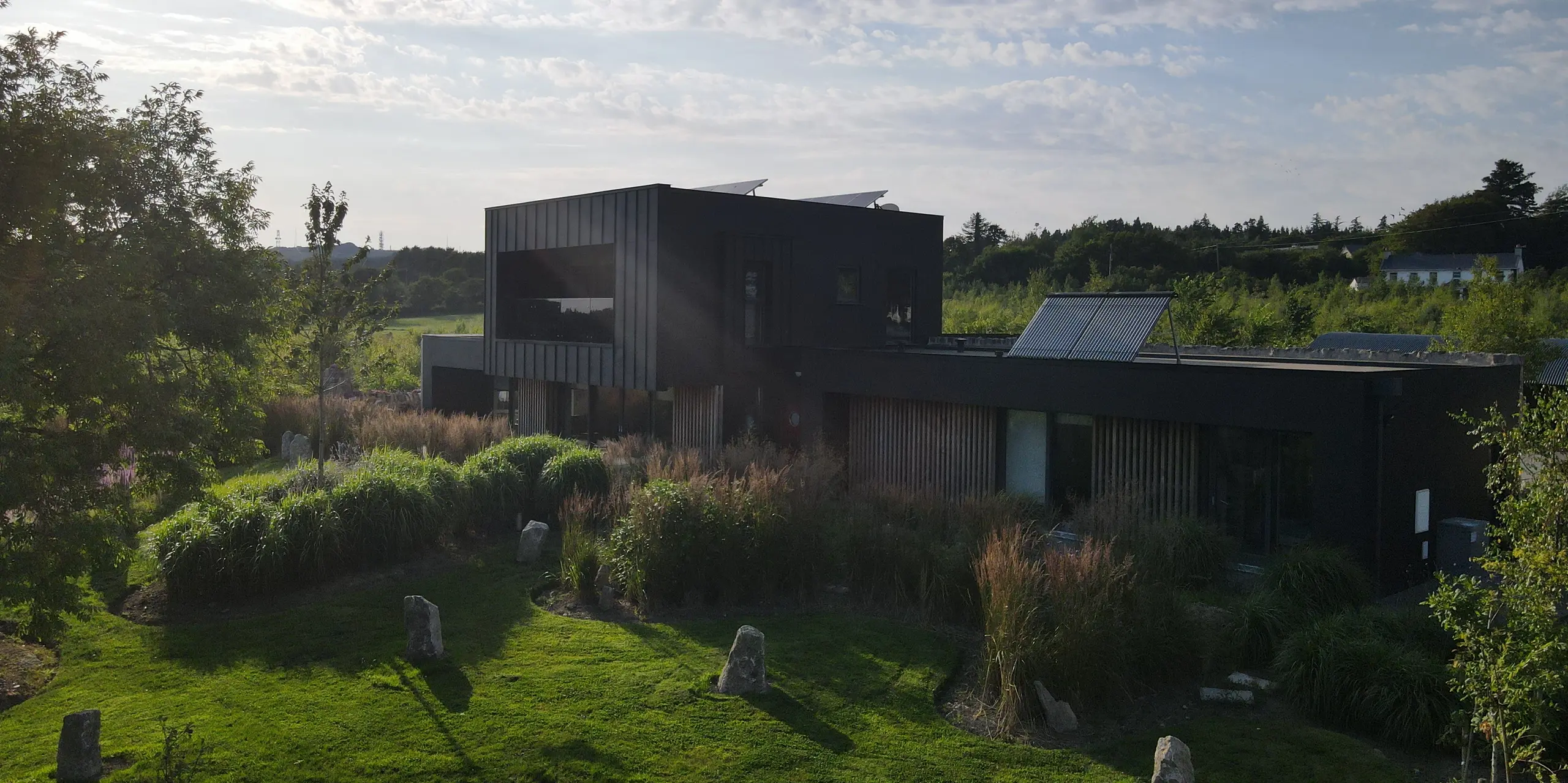 La photographie présente une maison individuelle moderne à Wexford, nichée au cœur d'un jardin luxuriant et paysagé. La façade noire PREFALZ, coloris P.10 noir, est particulièrement saisissante : ses lignes épurées et son élégant aspect métallique contrastent avec la verdure éclatante. La lumière intense du soleil couchant accentue la texture du revêtement en aluminium et souligne ses propriétés de haute qualité et sa résistance aux intempéries. L'alliance architecturale de formes cubiques modernes, de grandes fenêtres et du matériau PREFA robuste fait de ce bâtiment un véritable emblème architectural.
