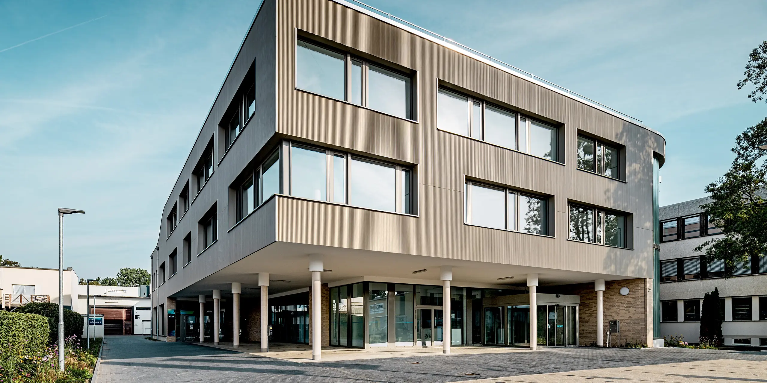L'image présente le bâtiment moderne Festo à Langenhagen, dont l'impressionnante façade se distingue visuellement grâce aux produits PREFA en bronze. La structure claire des profilés extrudés triangle confère à la façade une texture dynamique qui, associée aux élégants panneaux composites en aluminium du côté opposé, crée un ensemble harmonieux. La courbe remarquable de l'angle du bâtiment est particulièrement visible, apportant à la structure une élégante légèreté. Grâce aux matériaux PREFA, la façade est non seulement esthétique, mais aussi durable, facile d'entretien et résistante aux intempéries – des atouts essentiels pour un bâtiment pérenne.