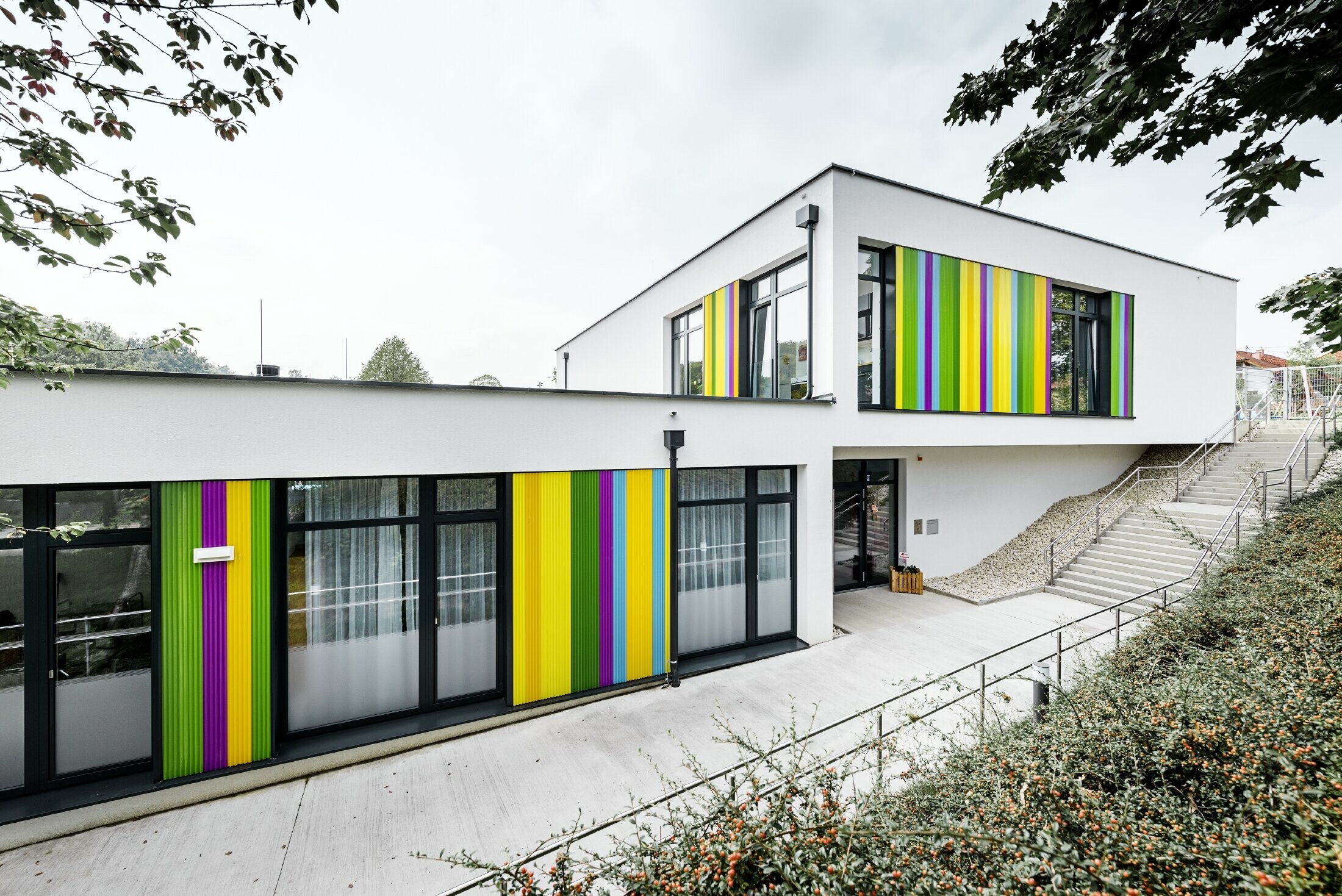 Éléments de façade multicolores de PREFA pour le jardin d’enfants de Hargelsberg. Le bâtiment présente un toit plat et des grandes fenêtres au niveau du sol.