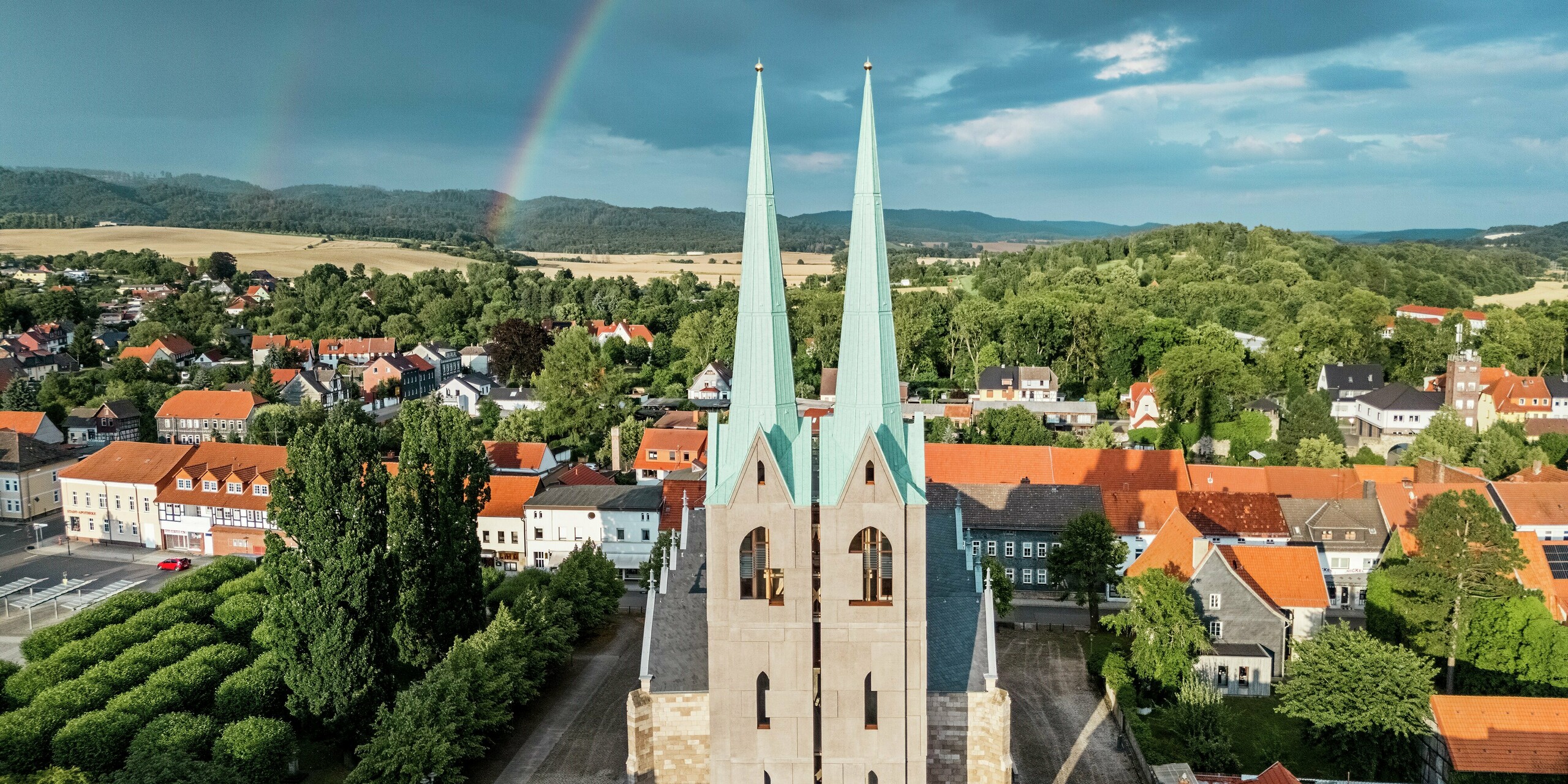 Beeindruckende Frontansicht des Tourismuszentrums Ellrich mit markanten Turmspitzen aus PREFALZ in P.10 Patinagrün – vor dramatischem Himmel und einem leuchtenden Regenbogen. Die Aluminiumdächer strahlen trotz dunkler Wolken und zeigen die Widerstandsfähigkeit und Ästhetik von PREFALZ selbst bei wechselhaftem Wetter. Ein Highlight moderner Sanierungskunst.