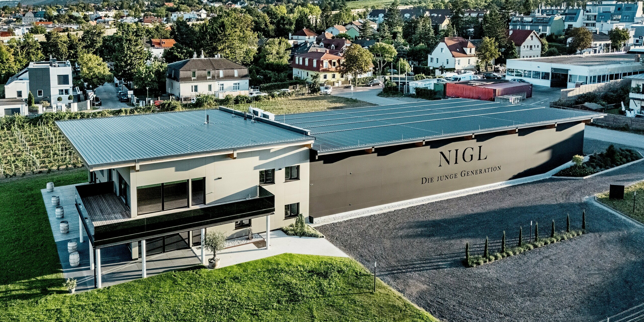 Luftaufnahme des Weinguts Nigl in Perchtoldsdorf mit moderner PREFA Aluminiumfassade in den Farben P.10 Schwarz und P.10 Prefabronze. Durch seine klare, kubische Architektur und den harmonischen Materialmix hebt sich das Gebäude deutlich von der umgebenden Wohn- und Weinlandschaft ab. Besonders markant ist die großflächige Seitenfassade mit dem Schriftzug „NIGL – Die junge Generation“. Das Flachdach mit sanftem Gefälle und die großzügige Terrasse mit Glasbrüstung unterstreichen den zeitgemäßen Charakter des Neubaus. Die PREFA Fassadenplatten überzeugen nicht nur durch ihre ästhetische Wirkung, sondern auch durch Langlebigkeit, Stabilität und optimalen Schutz vor Witterungseinflüssen.