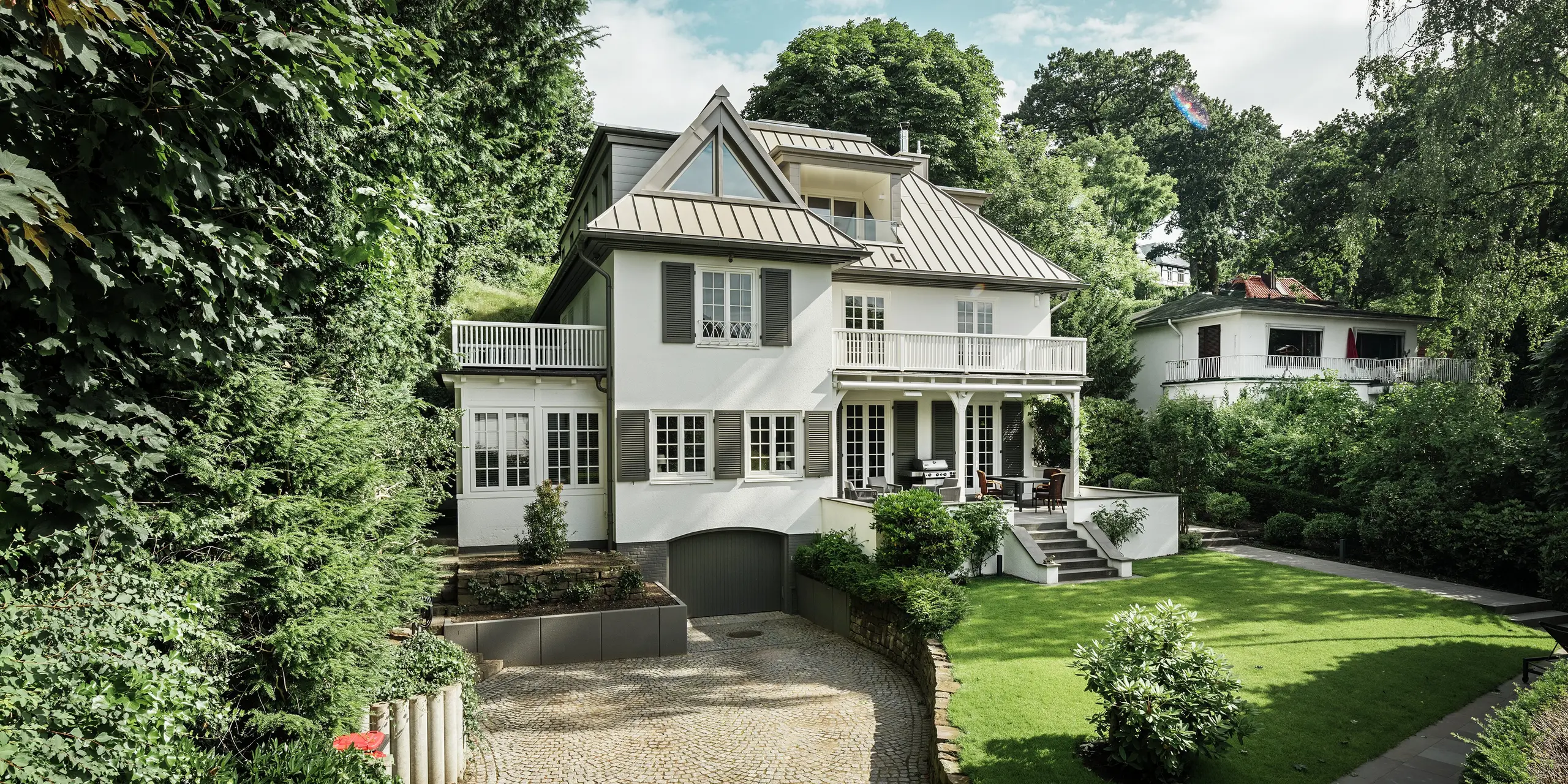 Das Bild zeigt die Villa Blankenese in Hamburg mit einem großzügigen Vorgarten und einer eleganten Zufahrt. Das markante Dach aus PREFALZ in Bronze prägt die Architektur und sorgt für eine klare, moderne Linienführung. Die Stehfalztechnik bietet nicht nur ein edles Erscheinungsbild, sondern auch höchste Witterungsbeständigkeit. Ergänzend dazu sind an den Gauben und Fassadenteilen bronzefarbene PREFA Sidings eingesetzt, die das harmonische Zusammenspiel von Dach und Fassade unterstreichen. Die Kombination aus langlebigem Aluminium und klassischem Design macht die Villa zu einem stilvollen Referenzobjekt für anspruchsvolle Architektur.