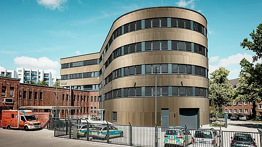 Vue extérieure saisissante du commissariat de police 43 à Hambourg : le nouveau bâtiment incurvé avec une façade en aluminium PREFA composée de profils extrudés anodisés « triangle » en bronze moyen constitue un puissant accroche-regard dans le paysage urbain. La structure fine et verticale crée des effets d'ombre et de lumière vifs et souligne l'architecture arrondie. Le stock de briques historique contraste avec cela. L'aluminium PREFA impressionne par sa haute résistance aux intempéries, sa protection contre la corrosion, sa longévité et son entretien minimal - idéal pour des solutions de façade durables, fonctionnelles et esthétiques.