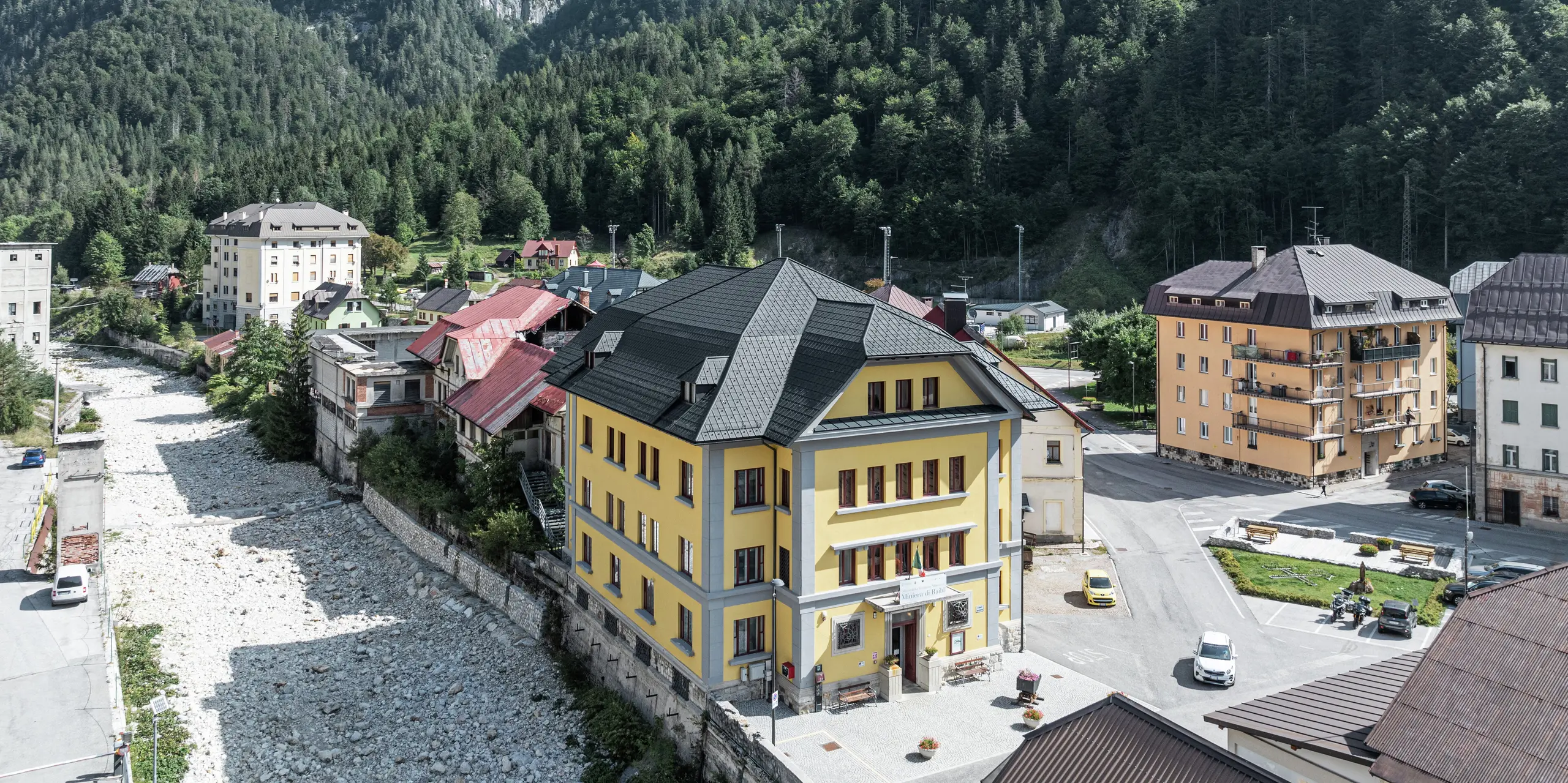 Niché dans un paysage de montagne impressionnant se trouve le Museo Cave del Predil en Italie. Le musée avec sa façade jaune frappante est équipé de tuiles PREFA de haute qualité de couleur P.10 anthracite. Le toit en aluminium durable garantit une protection optimale contre les conditions météorologiques alpines telles que la neige et les tempêtes, associée à un design esthétique. En arrière-plan, le panorama alpin s'élève majestueusement avec des forêts denses et des pics rocheux.