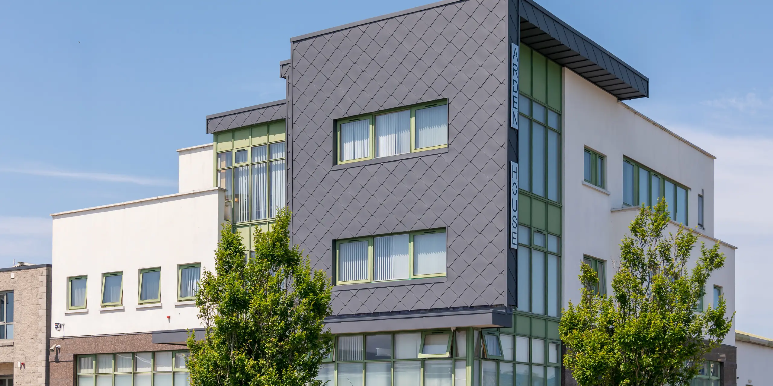 Vue oblique du centre de santé mentale pour enfants et jeunes de Wexford, mettant en valeur son remarquable revêtement de façade PREFA. L'attention se porte sur la partie cubique du bâtiment, habillée de losanges 44 x 44 en P.10 gris sombre, dont l'élégante structure en aluminium crée un jeu d'ombres et de lumières saisissant. La façade en losanges foncés contraste de façon captivante avec les surfaces claires et les larges baies vitrées. Durable, inaltérable et résistant aux intempéries, le système PREFA confère au bâtiment un style architectural moderne et de grande qualité, empreint d'un caractère distinctif.
