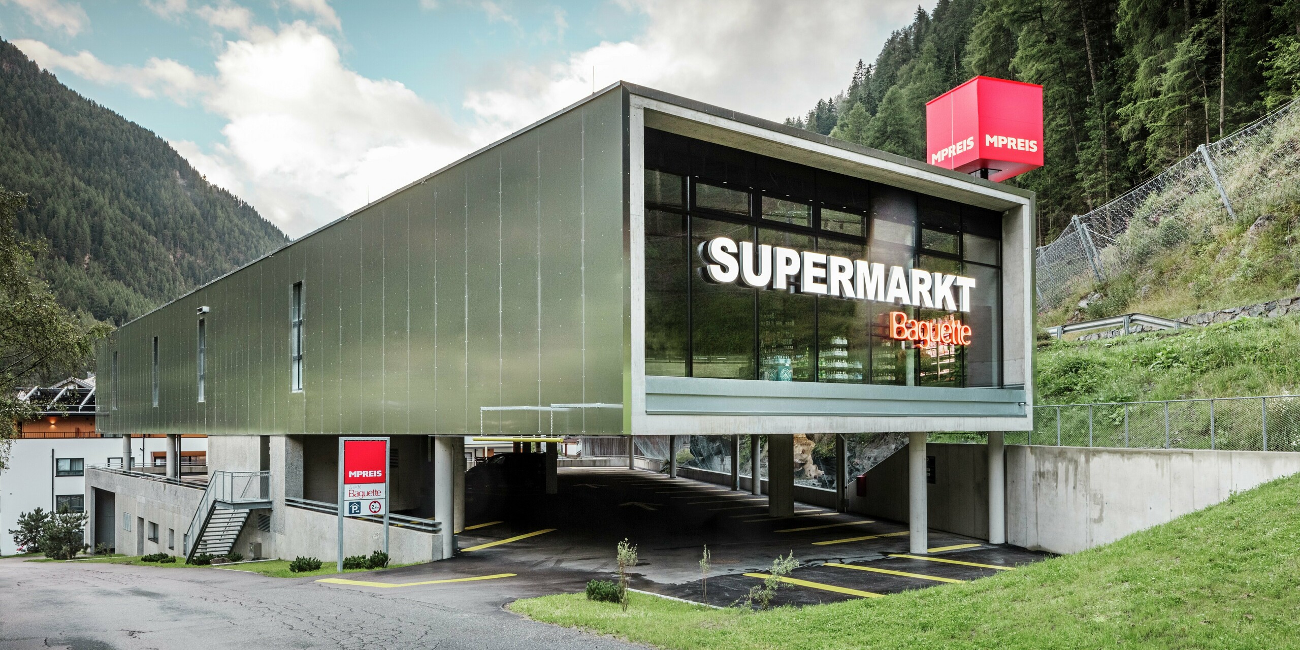 Accès au supermarché MPREIS à Sölden, au Tyrol, avec des panneaux composites en aluminium dans la couleur spéciale Reflective Natural. La façade moderne reflète les montagnes environnantes. De généreuses façades vitrées, un parking surélevé et des lignes claires allient fonctionnalité et design contemporain dans la région alpine.