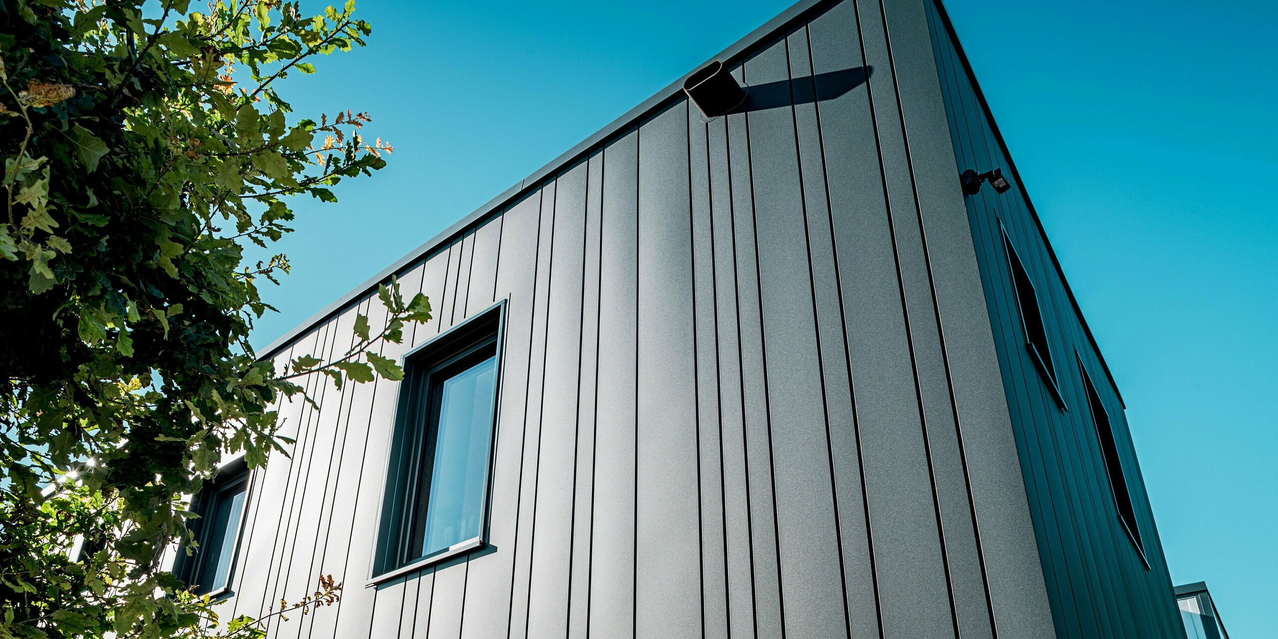 Der Blick auf die Gebäudeecke zeigt eindrucksvoll die präzise Verarbeitung der PREFA Sidings. Die vertikale Schattenfuge erzeugt Tiefe und eine klare Linienästhetik. Im Sonnenlicht schimmern die hochwertig beschichteten Aluminiumoberflächen in kräftigem Anthrazit. Diese Detailansicht unterstreicht die Langlebigkeit, Formstabilität und optische Eleganz der PREFA Fassadensysteme.