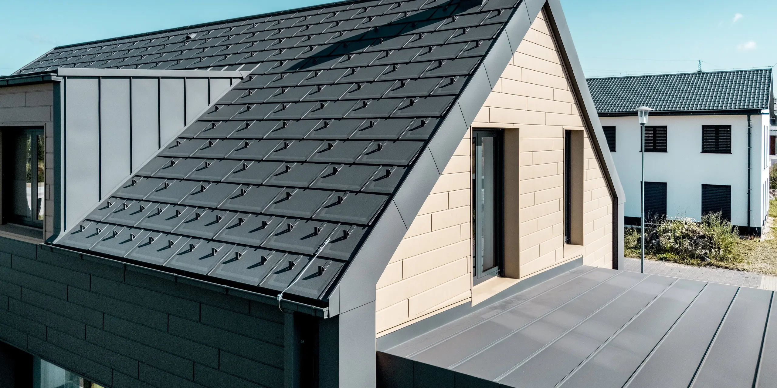 Detailaufnahme des modernen Einfamilienhauses mit PREFA Dachplatte R.16 und PREFALZ in P.10 Dunkelgrau. Die klaren Linien der Aluminiumdächer und die sandbraune Siding-Fassade verbinden Ästhetik und Funktion. Die langlebigen PREFA Oberflächen bieten zuverlässigen Schutz vor Witterung und verleihen dem Gebäude eine zeitgemäße, harmonische Optik.