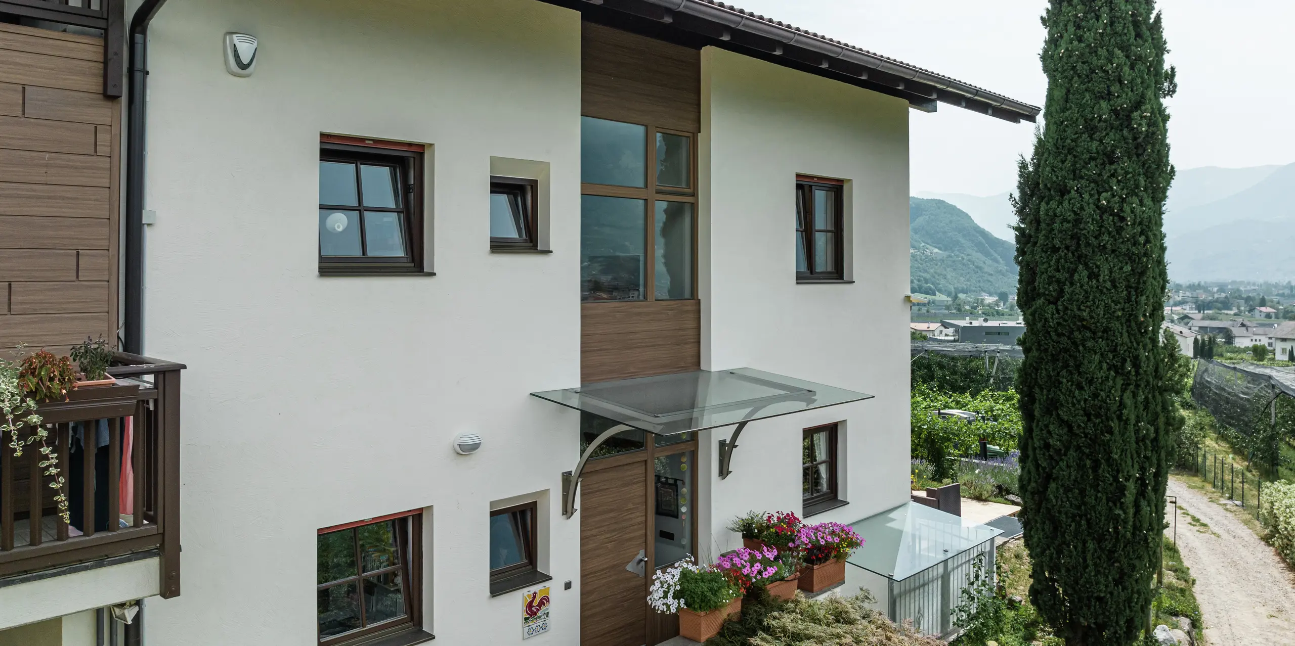 Zone d'entrée des appartements de vacances Prechtlhof à Merano, dans le Tyrol du Sud, avec une façade moderne en plâtre et des Sidings PREFA en noyer foncé. La façade en aluminium sans entretien avec un aspect bois complète le design traditionnel et garantit une solution durable et résistante aux intempéries.