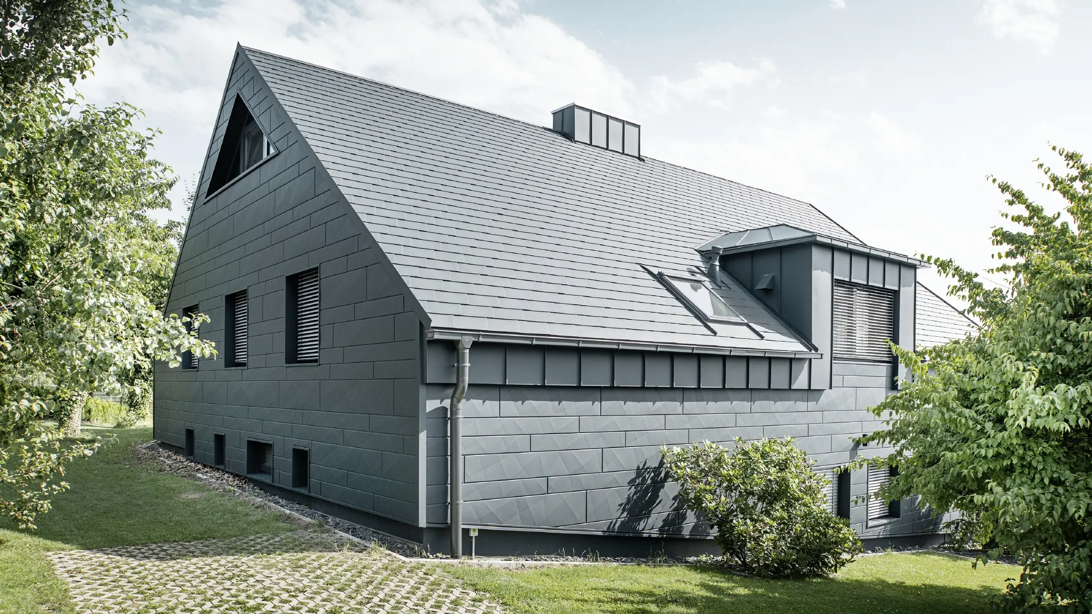 Grande maison individuelle avec toit en pente et nombreuses fenêtres. L'ensemble de la maison a été revêtu d'une enveloppe en aluminium anthracite, la toiture a été réalisée avec des bardeaux en aluminium PREFA et la façade avec du Siding.X anthracite.