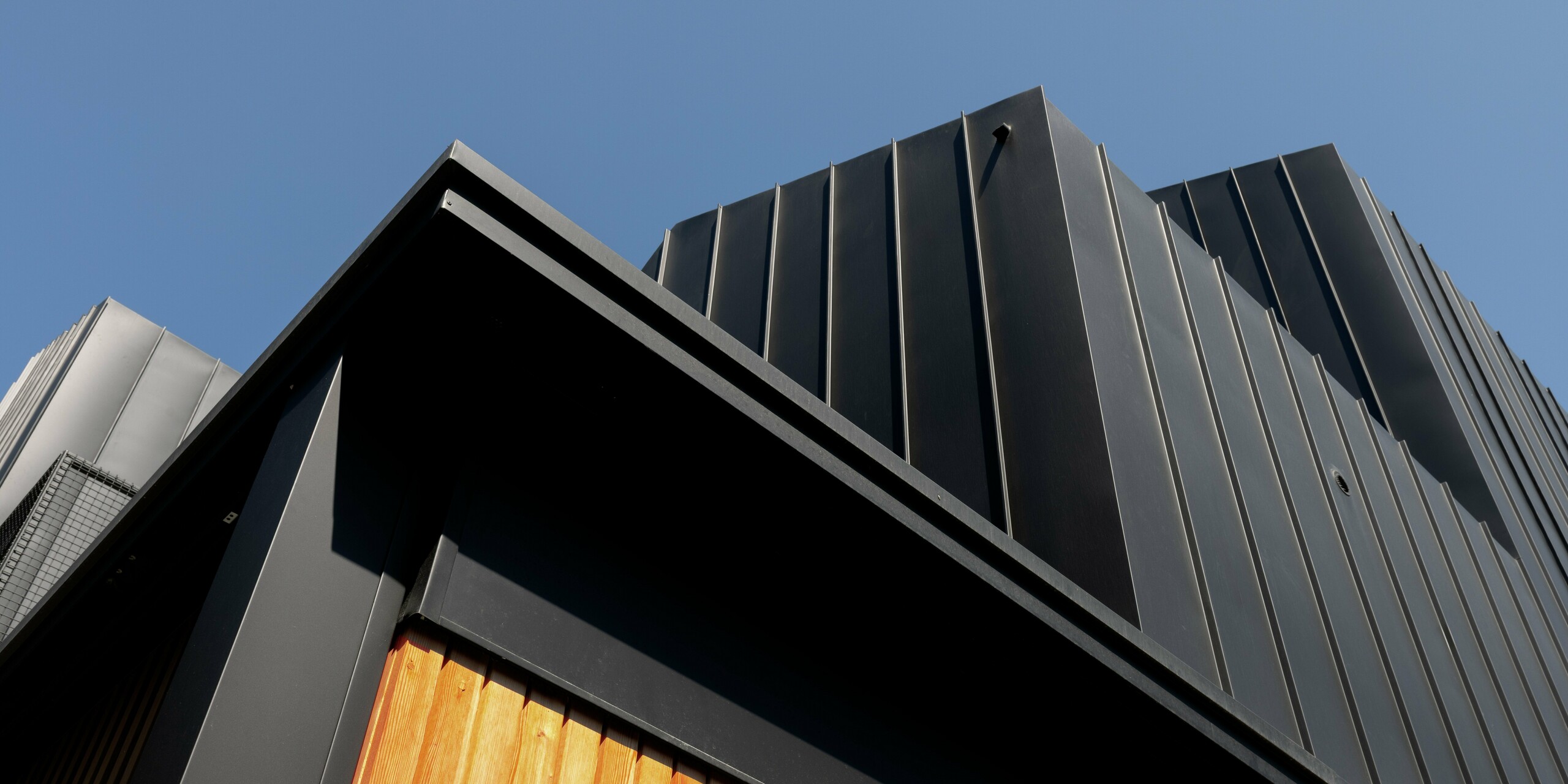 Nahaufnahme der modernen Architektur in Cattolica: markante Aluminiumfassade mit PREFALZ in Schwarzgrau, kombiniert mit warmer Holzverkleidung. Die klaren Linien und die hochwertige PREFA Oberfläche sorgen für Langlebigkeit, Korrosionsschutz und ein elegantes, zeitloses Design unter strahlend blauem Himmel.