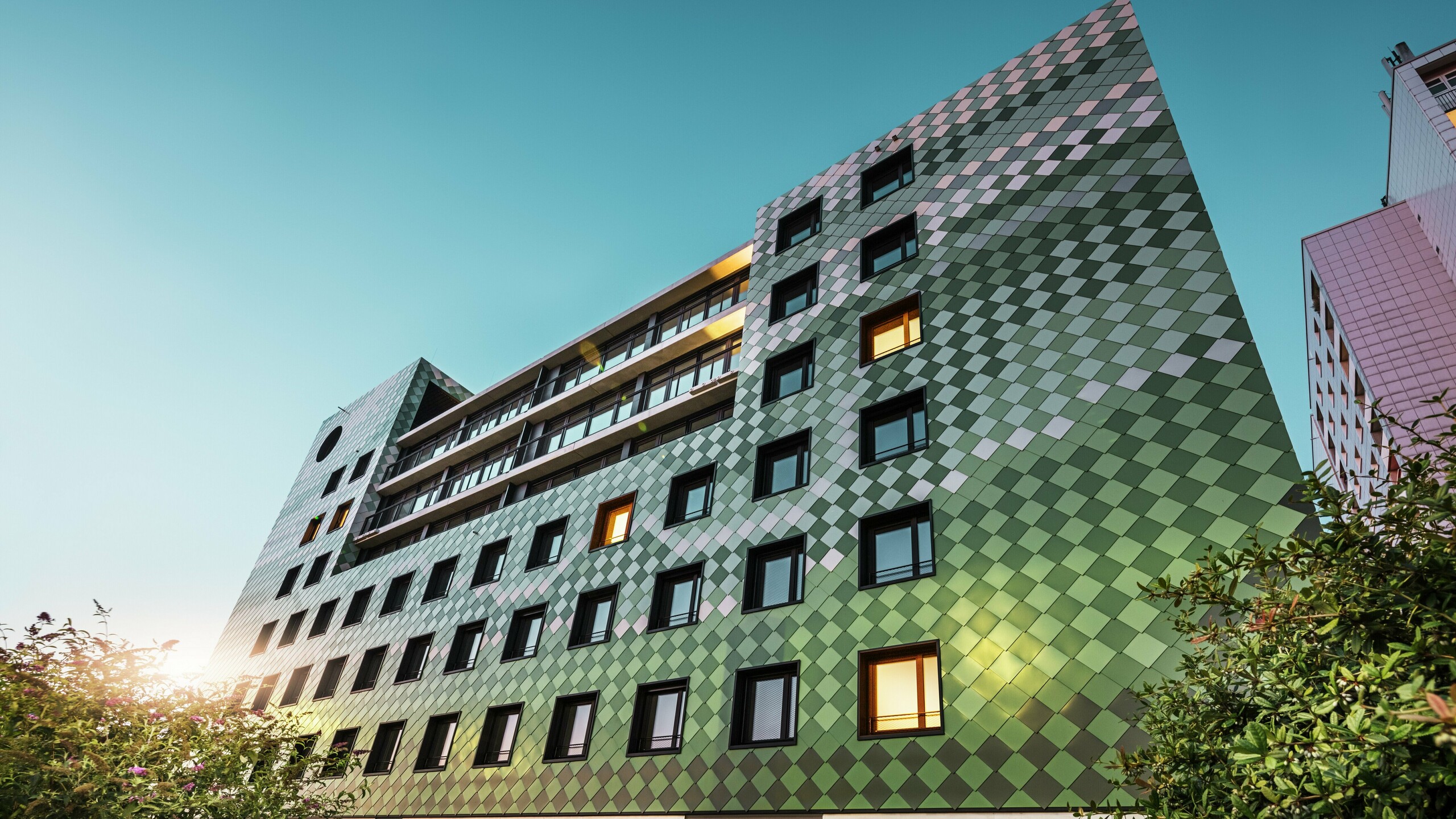Das moderne Wohn- und Geschäftsgebäude „Pick-Nick“ in Dresden besticht durch seine markante PREFA-Fassade aus Aluminium-Rhombusen in den Sonderfarben Olivgrün, Graugrün, Resedagrün, Opalgrün, Hellelfenbein, Perlweiß und Hellbronze. Die kreative Fassadengestaltung erinnert an ein Reisfeld und erzeugt ein lebendiges Farbenspiel. Die klare Architektur, die schwarzen Fensterrahmen und die Langlebigkeit der PREFA-Materialien prägen das Gebäude. Das tiefstehende Sonnenlicht spiegelt sich auf der Fassade und den Fensterflächen.